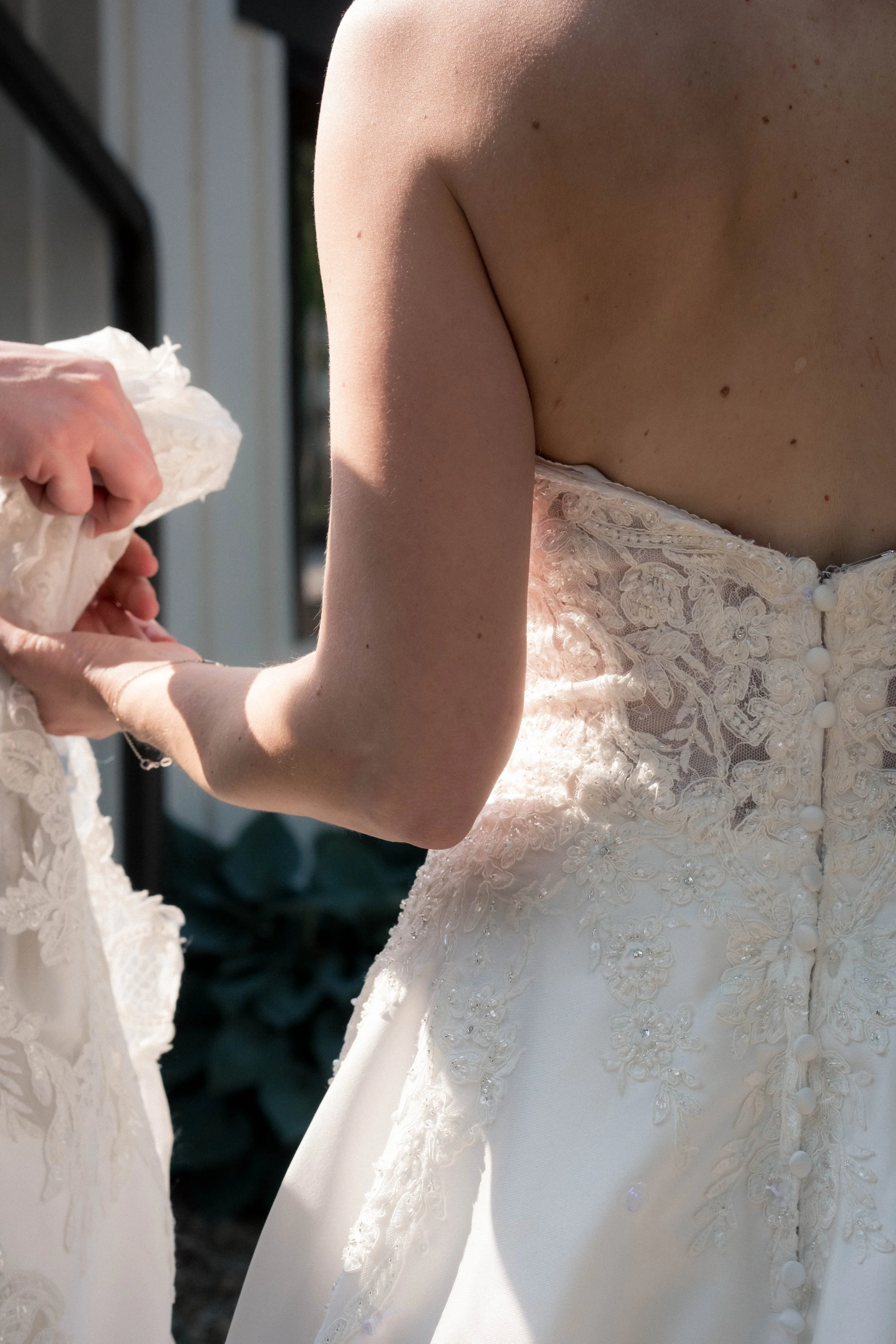 A person adjusting a wedding dress outdoors, showing the back of the dress with lace detailing and buttons.