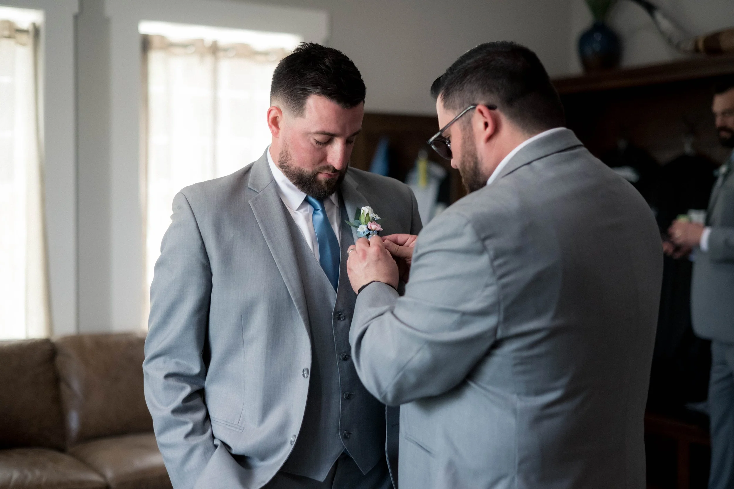 A man in a light gray suit has a blue tie, and another man in a similar suit pinning a boutonniere on his lapel during a wedding prep in a cozy room with sunlight and a brown couch.