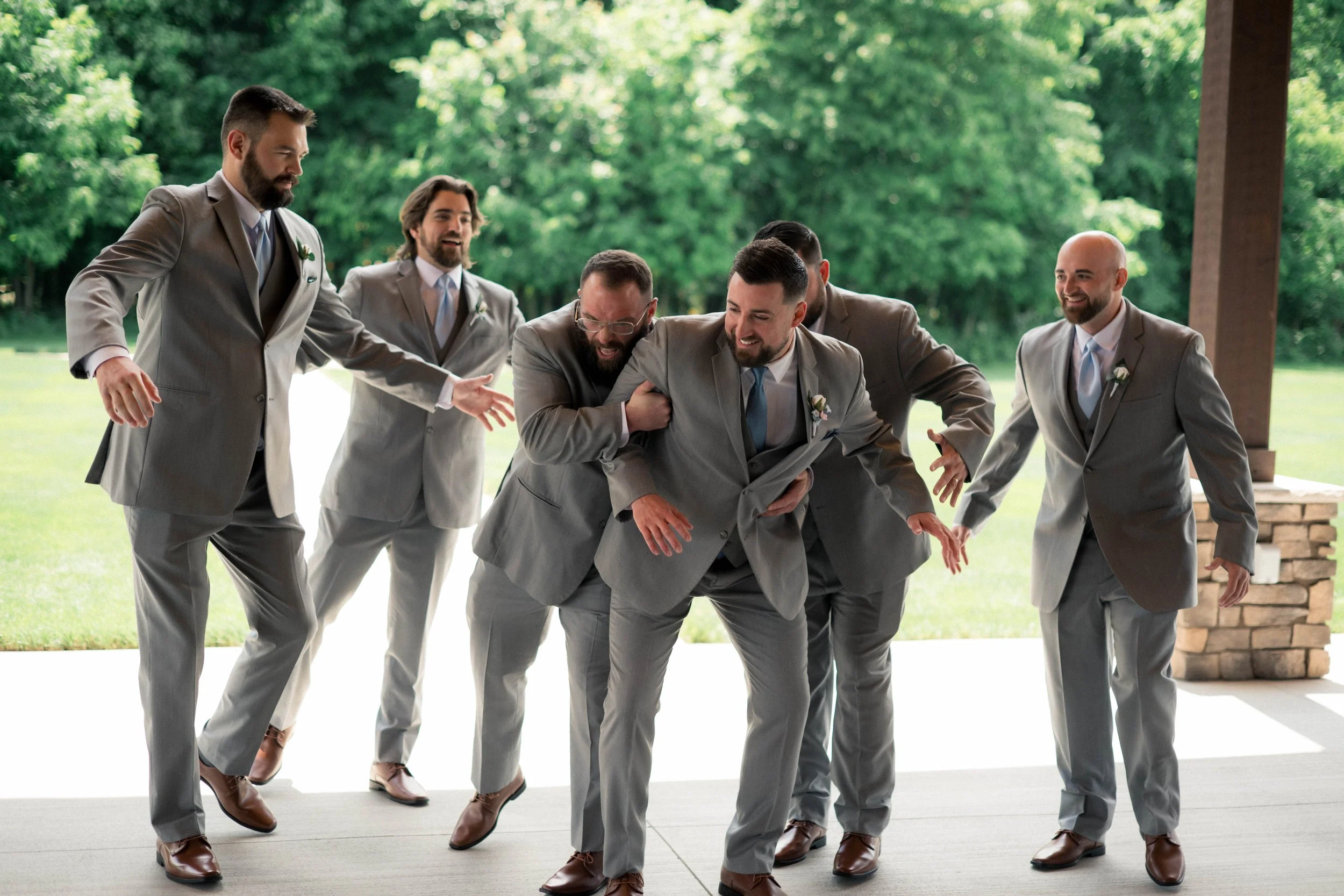 Groom and groomsmen dressed in gray suits having fun and playfully wrestling outdoors.