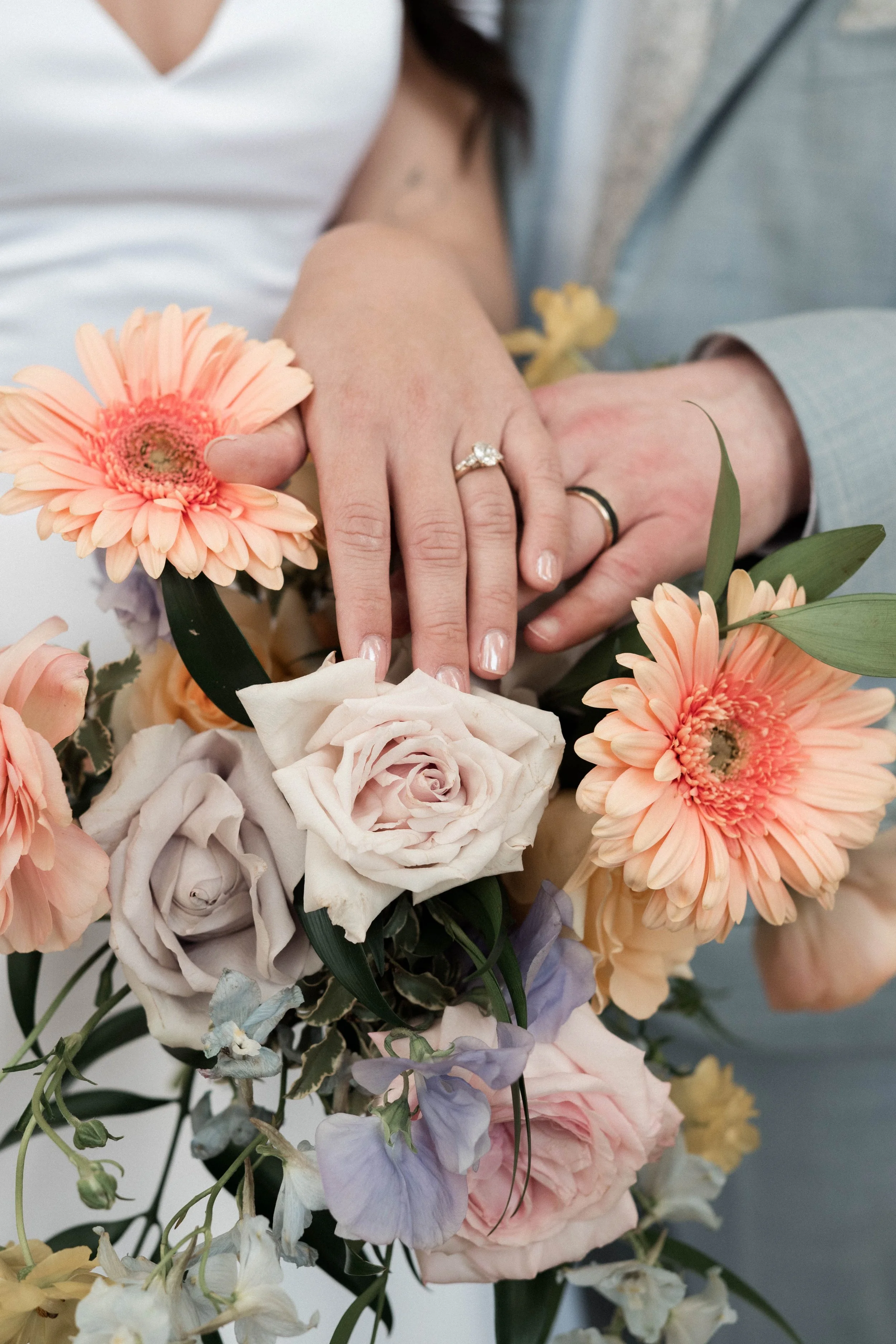 Close-up of a couple's hands showing wedding rings, resting on a bouquet of pastel-colored flowers including roses and gerbera daisies.