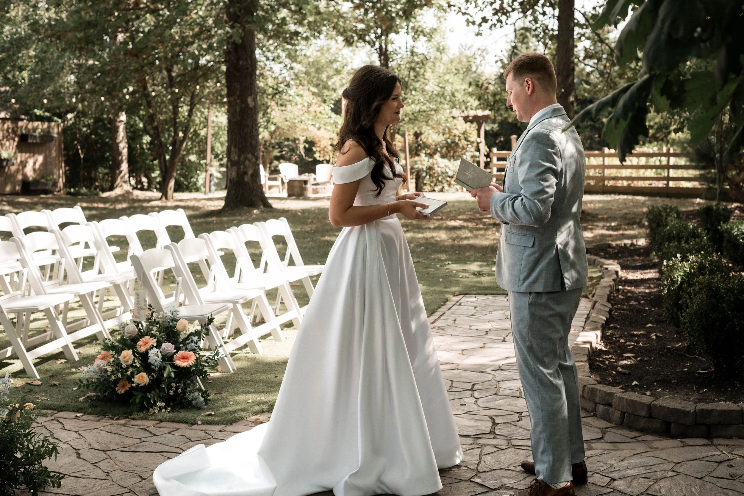 A bride and groom exchanging vows during outdoor wedding ceremony, surrounded by trees and white folding chairs, with a floral arrangement on the ground.