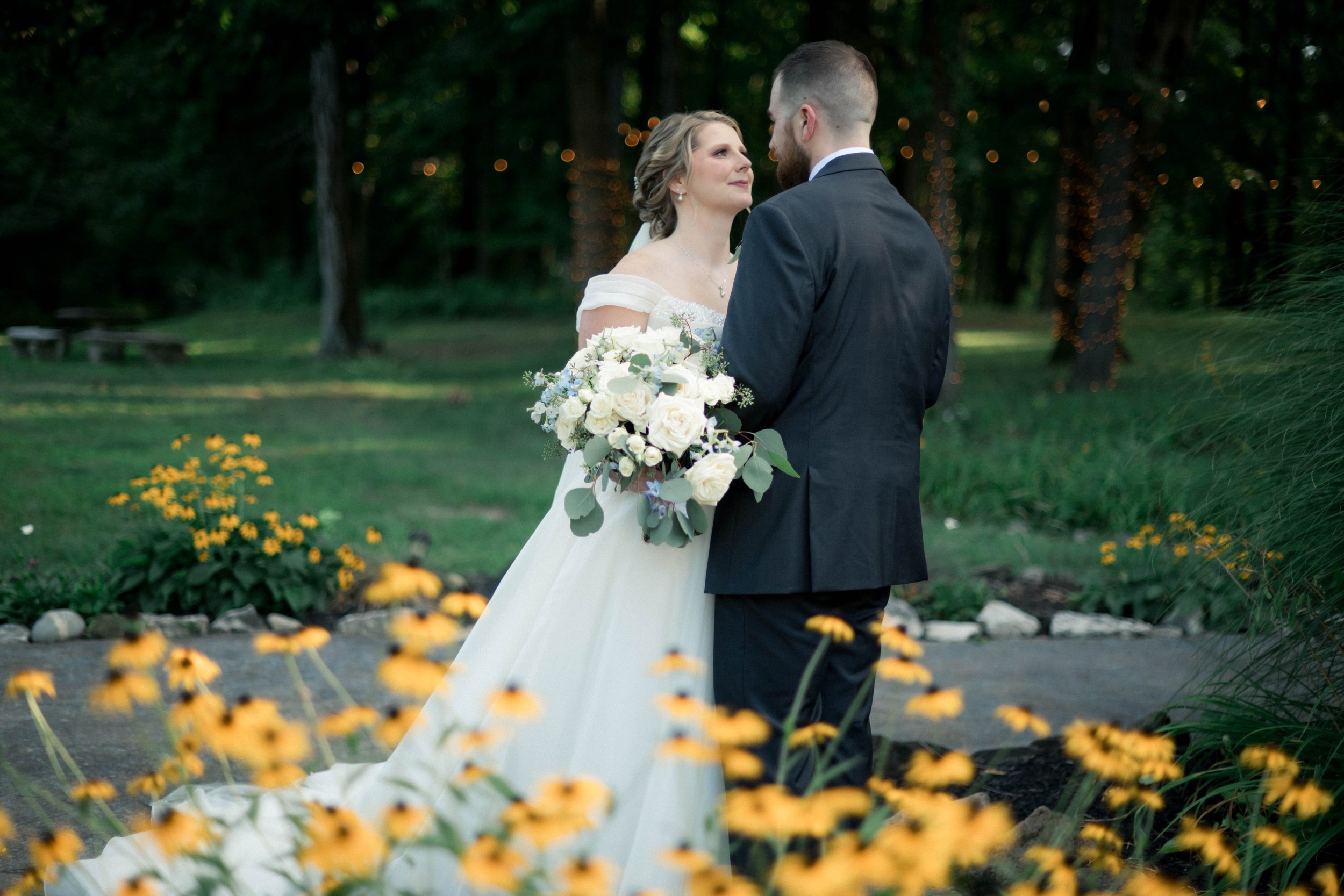 A bride and groom stand in a field of bright yellow flowers, gazing lovingly at one another on their wedding day in Columbus, OH. A candid moment captured by their photographer.