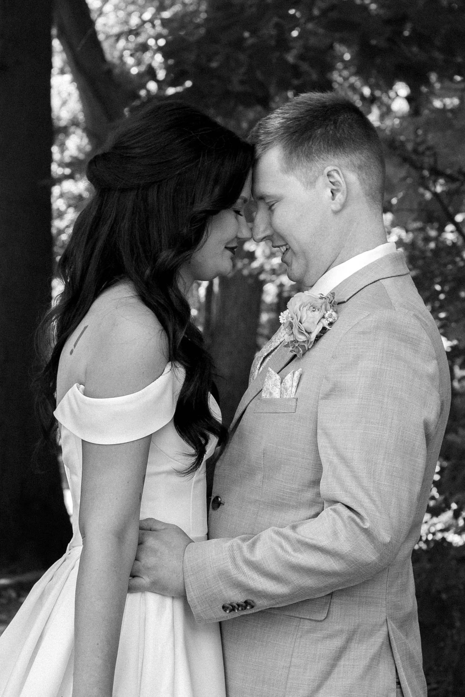 Black and white photo of a bride and groom with foreheads touching, smiling, outdoors surrounded by trees.