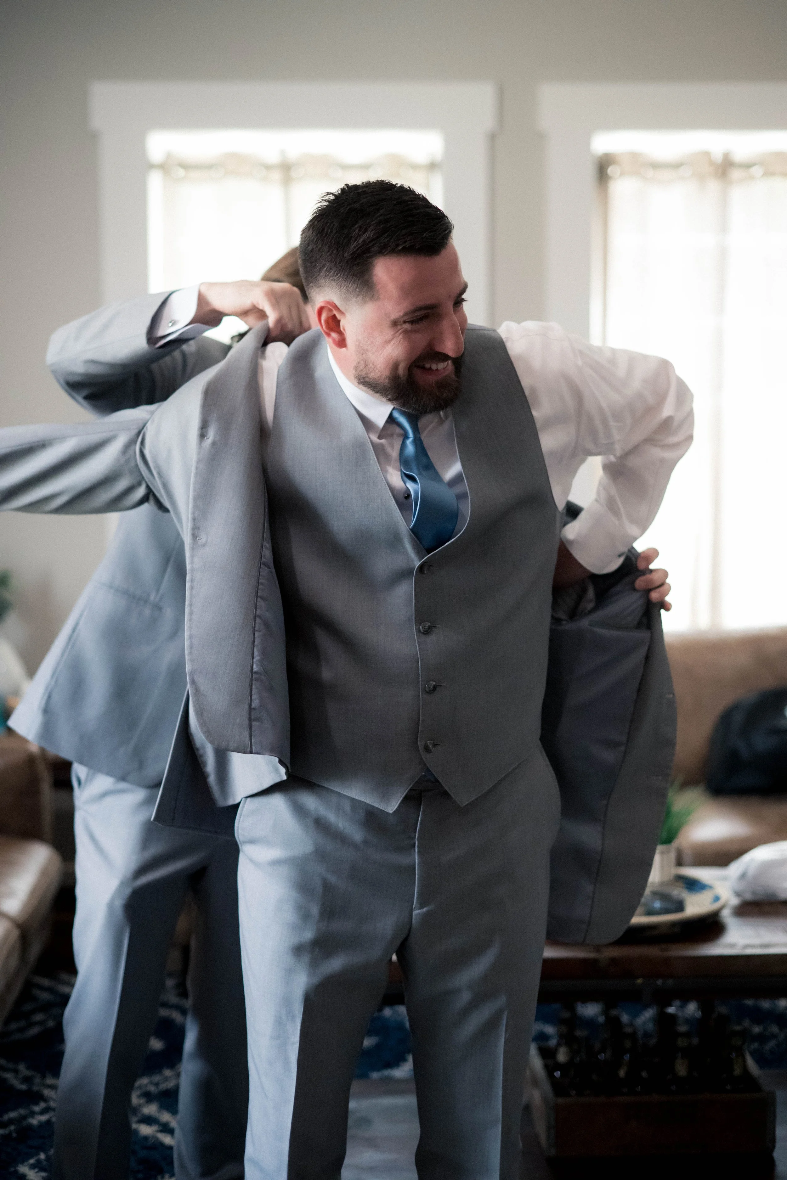 A man in a light gray suit, white shirt, and blue tie, smiling as he puts on his jacket with assistance from others in a well-lit room.