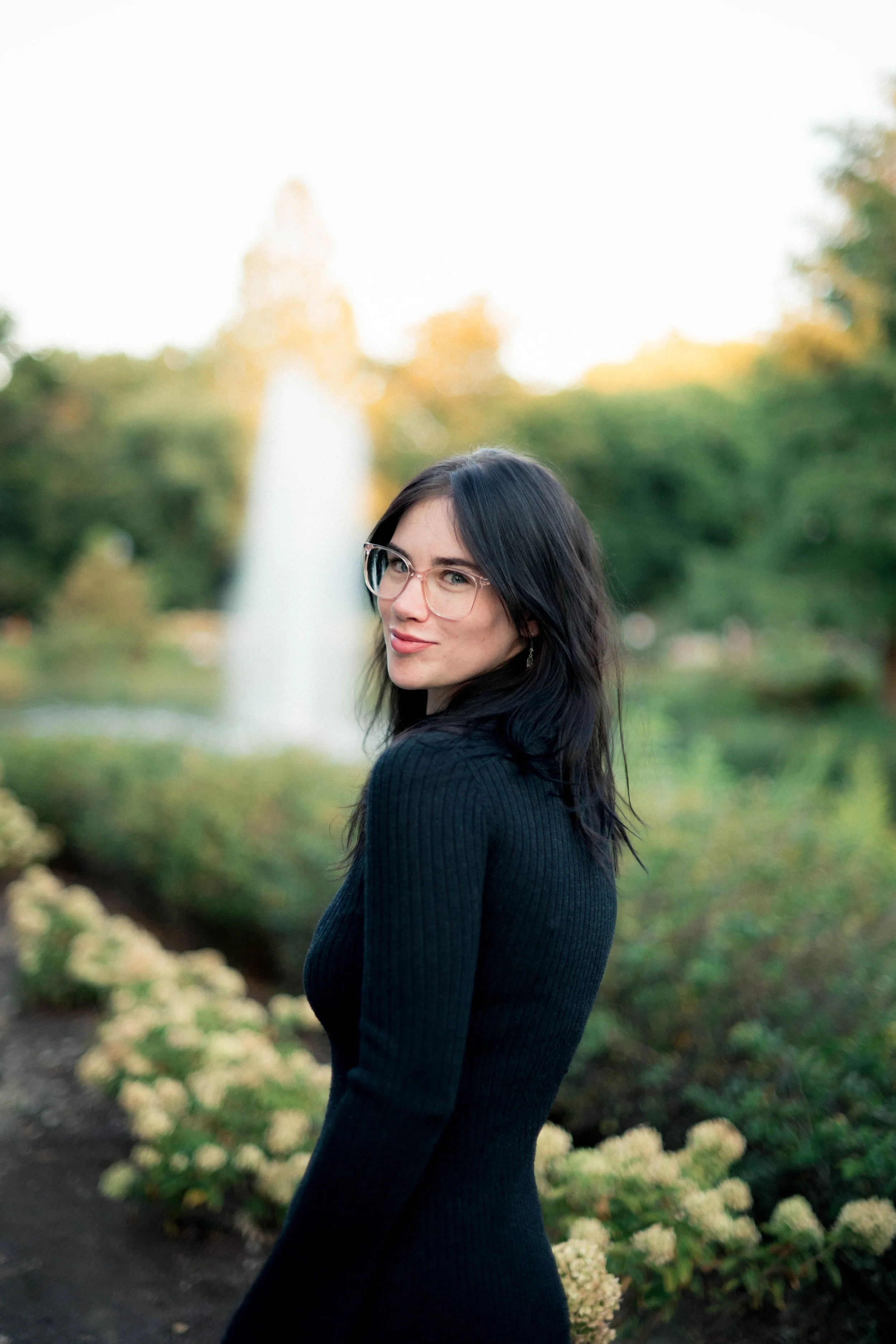 A woman with black hair, glasses, and a black long-sleeve dress standing outdoors near a garden with white flowers, with a fountain and green trees in the background.