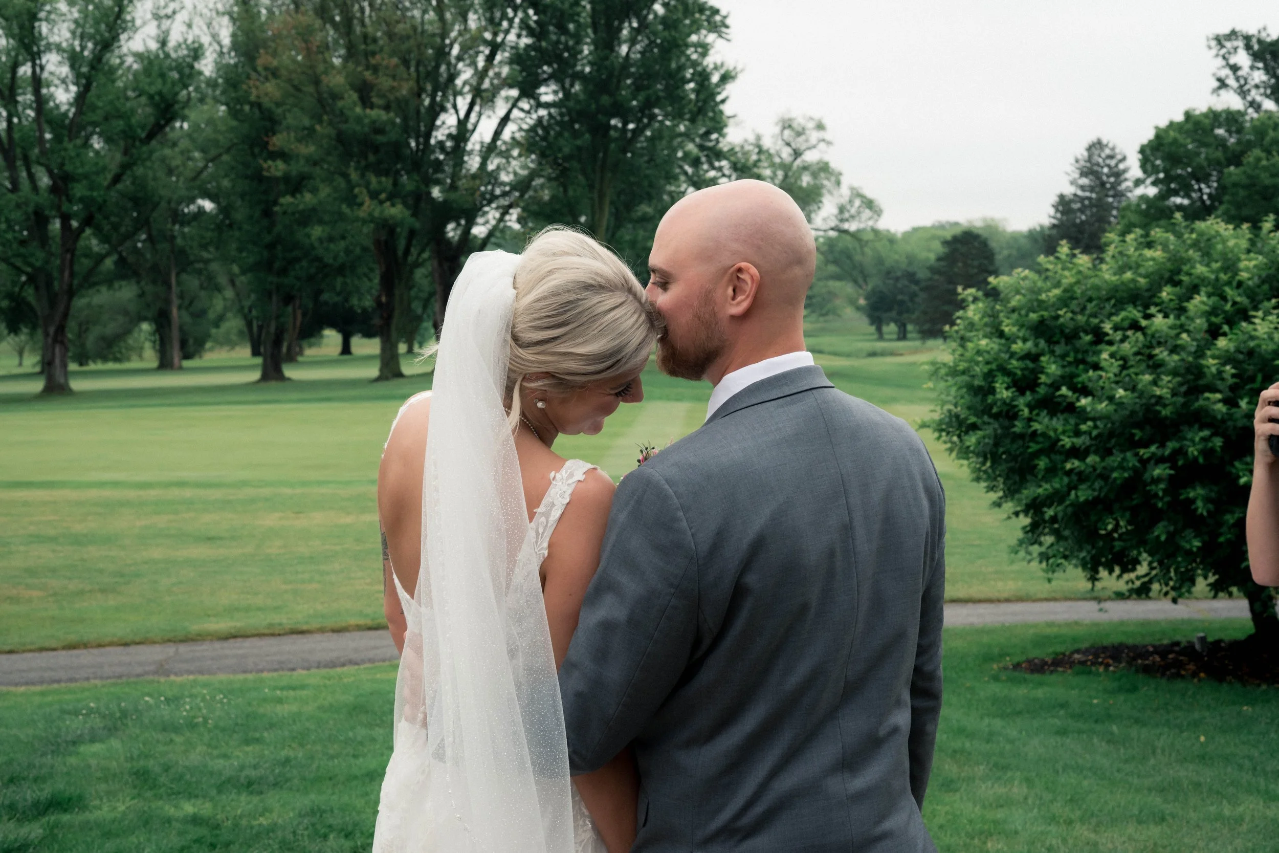 A bride and groom sharing a tender moment outdoors on their wedding day, with the groom kissing the bride on her forehead. They are surrounded by a lush green park with trees in the background.