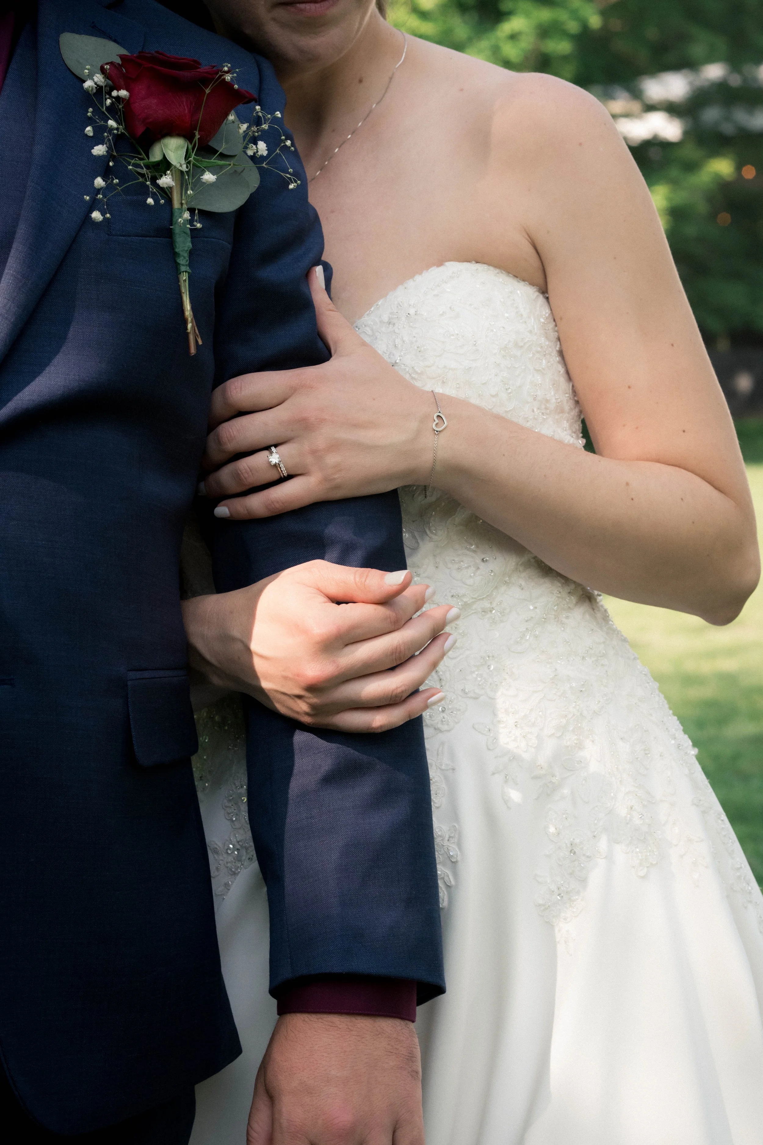 Close-up of a bride and groom holding each other outdoors, with focus on their intertwined hands, wedding dress, and suit jacket.