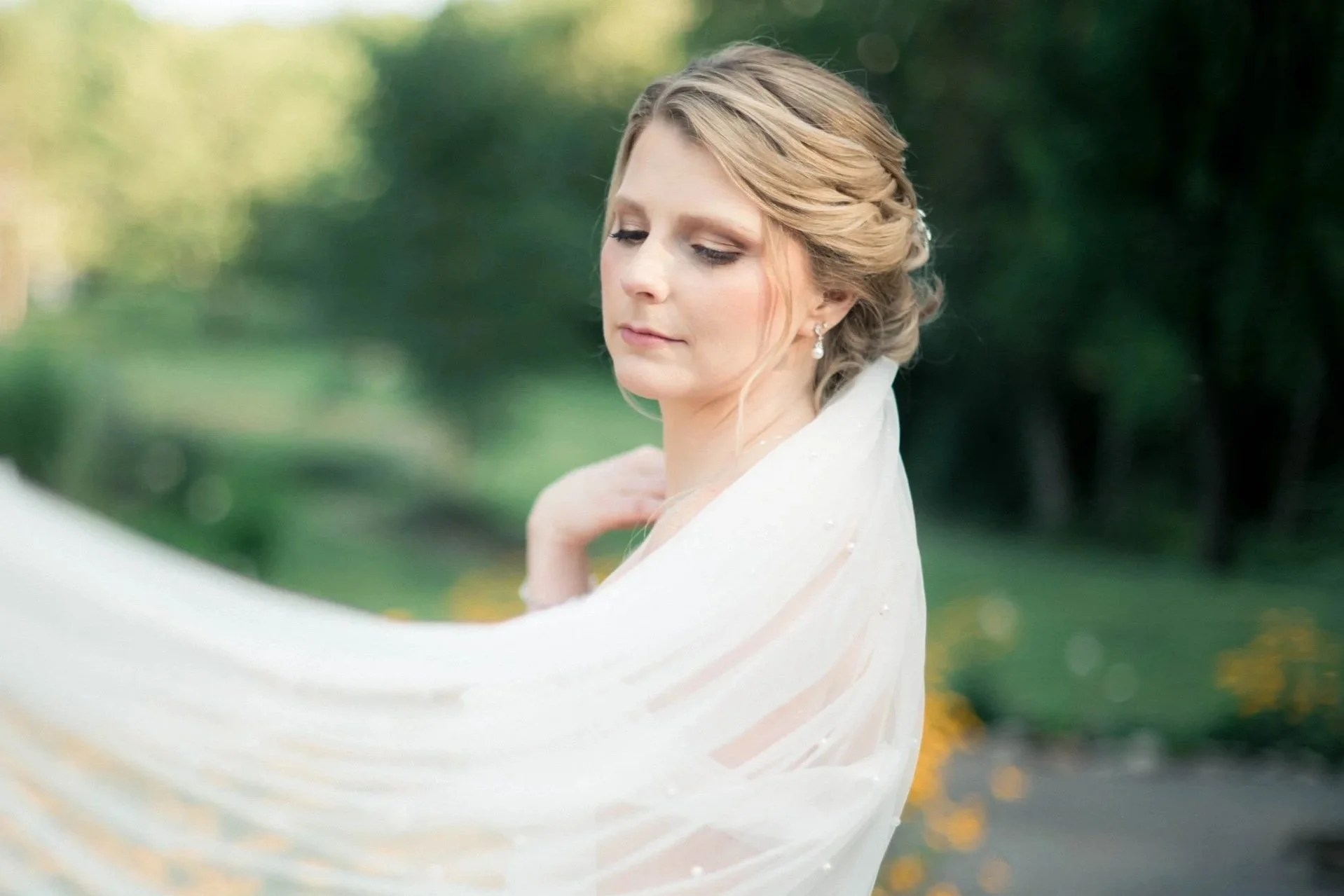 A bride in a white wedding dress standing outdoors with a lush green background, gently holding her wedding veil.