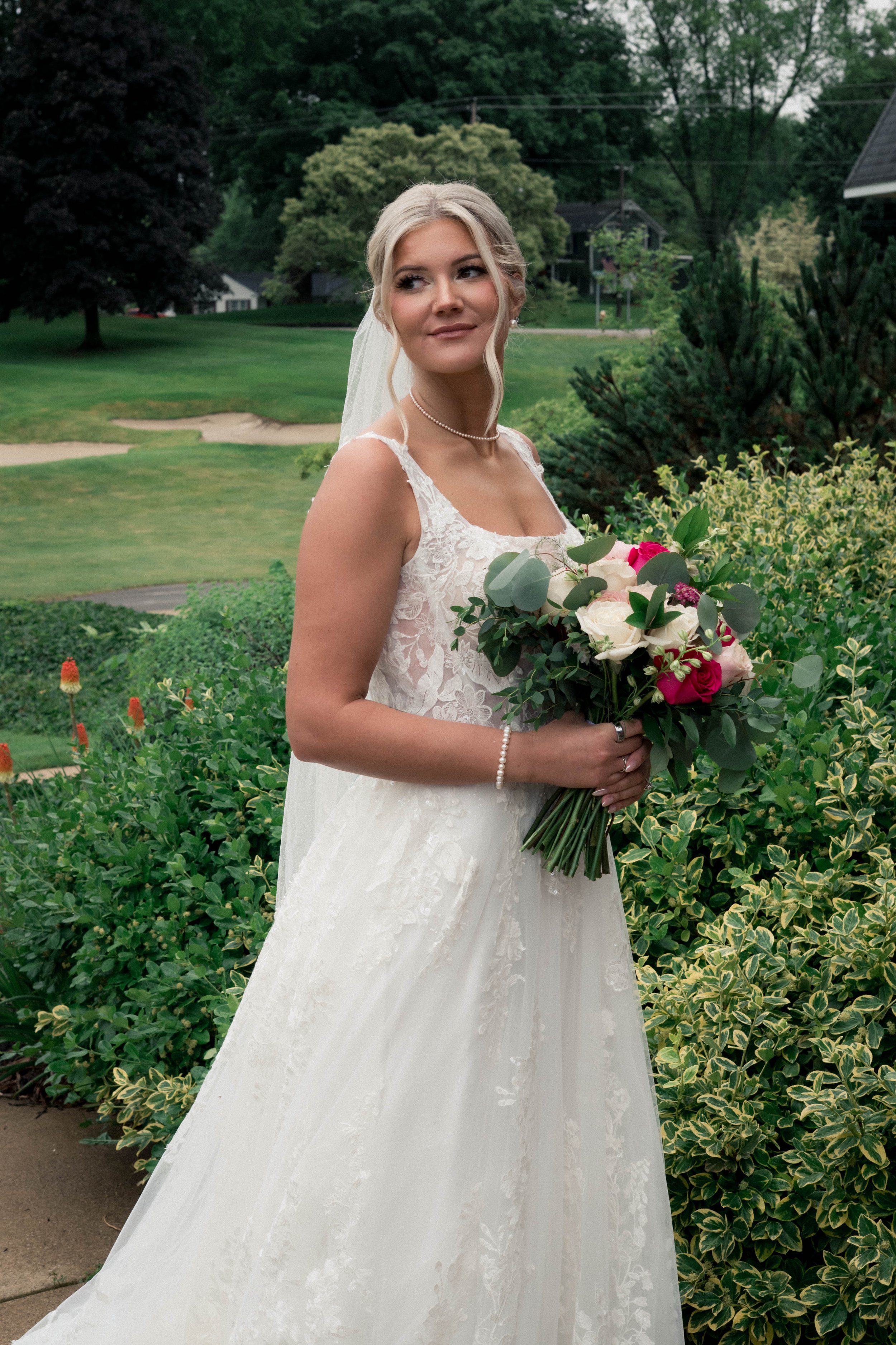 A bride in a wedding dress holding a bouquet of white and pink roses, standing outdoors among greenery, with trees in the background.