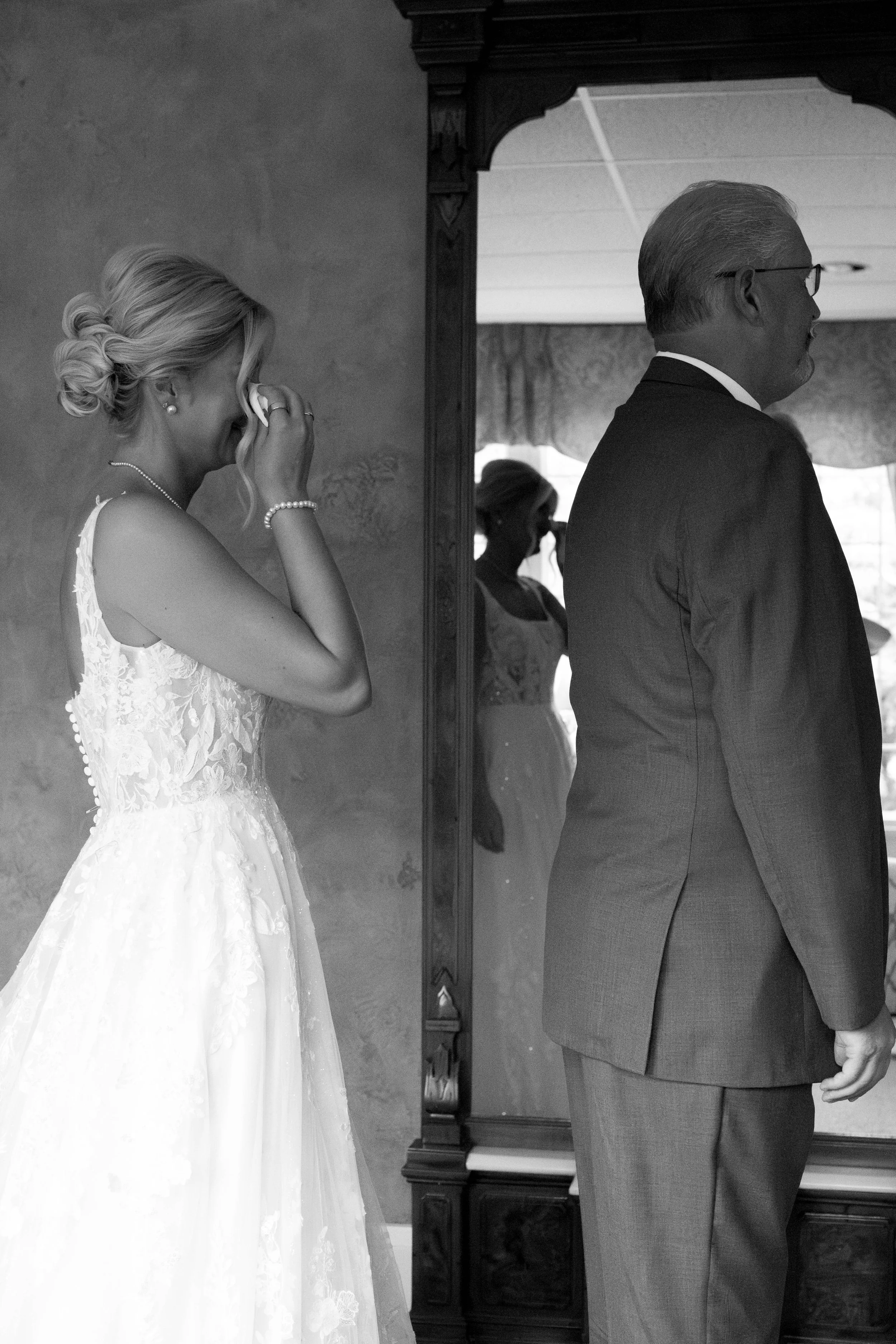 A bride wiping her eyes while standing next to a man in a suit, with a reflection of a woman holding a phone in a mirror in the background.