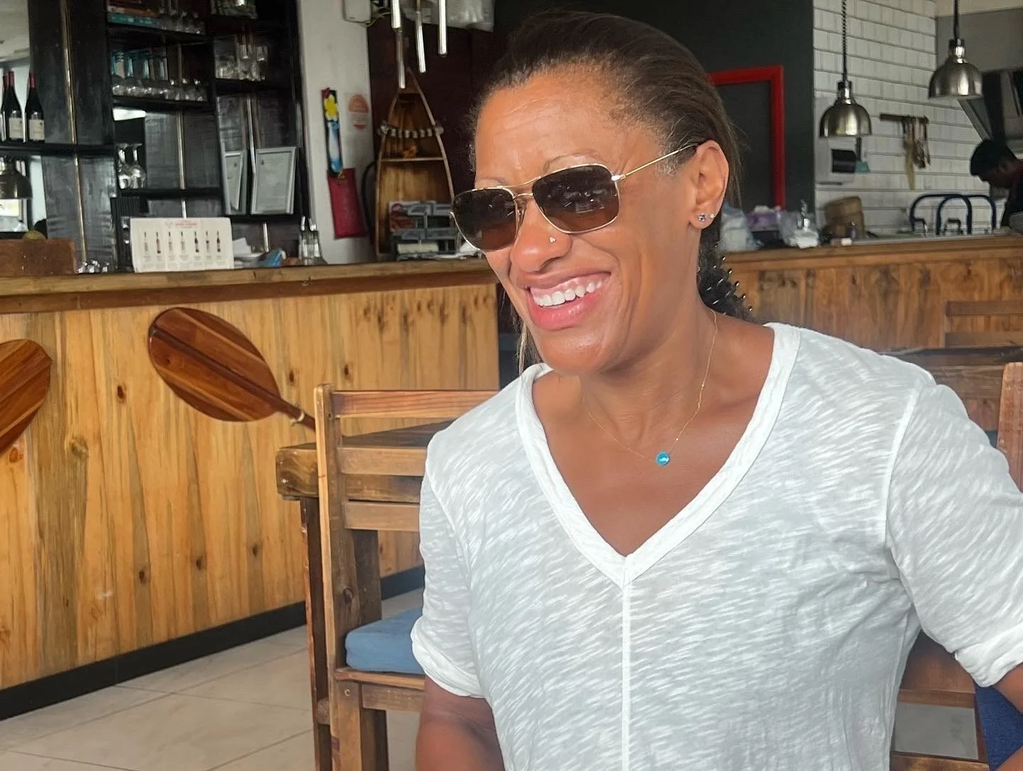 Person wearing sunglasses and a white shirt, sitting in a restaurant with wooden decor and paddles on the wall.