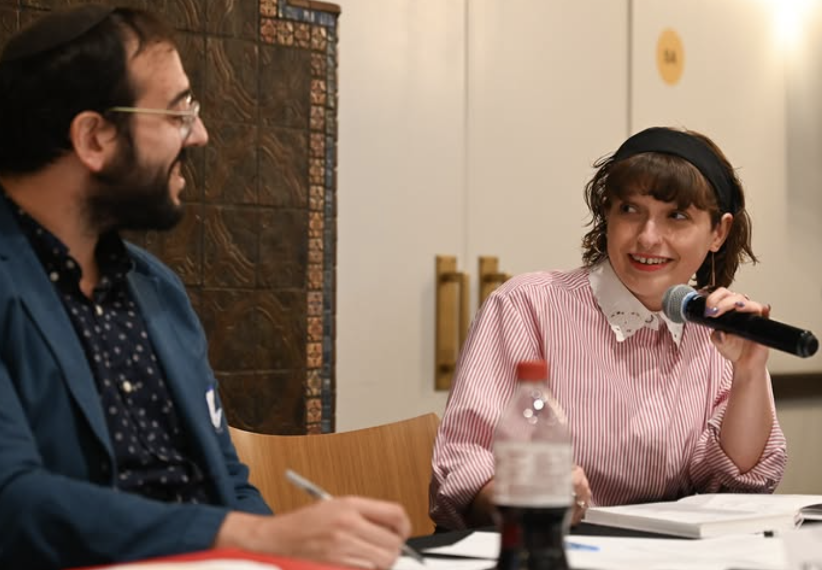 Two rabbis sitting at a table, engaging in conversation. The woman with short curly hair and a headband is holding a microphone and smiling, looking at the man with glasses and a beard. There are notebooks, a pen, and a water bottle on the table.