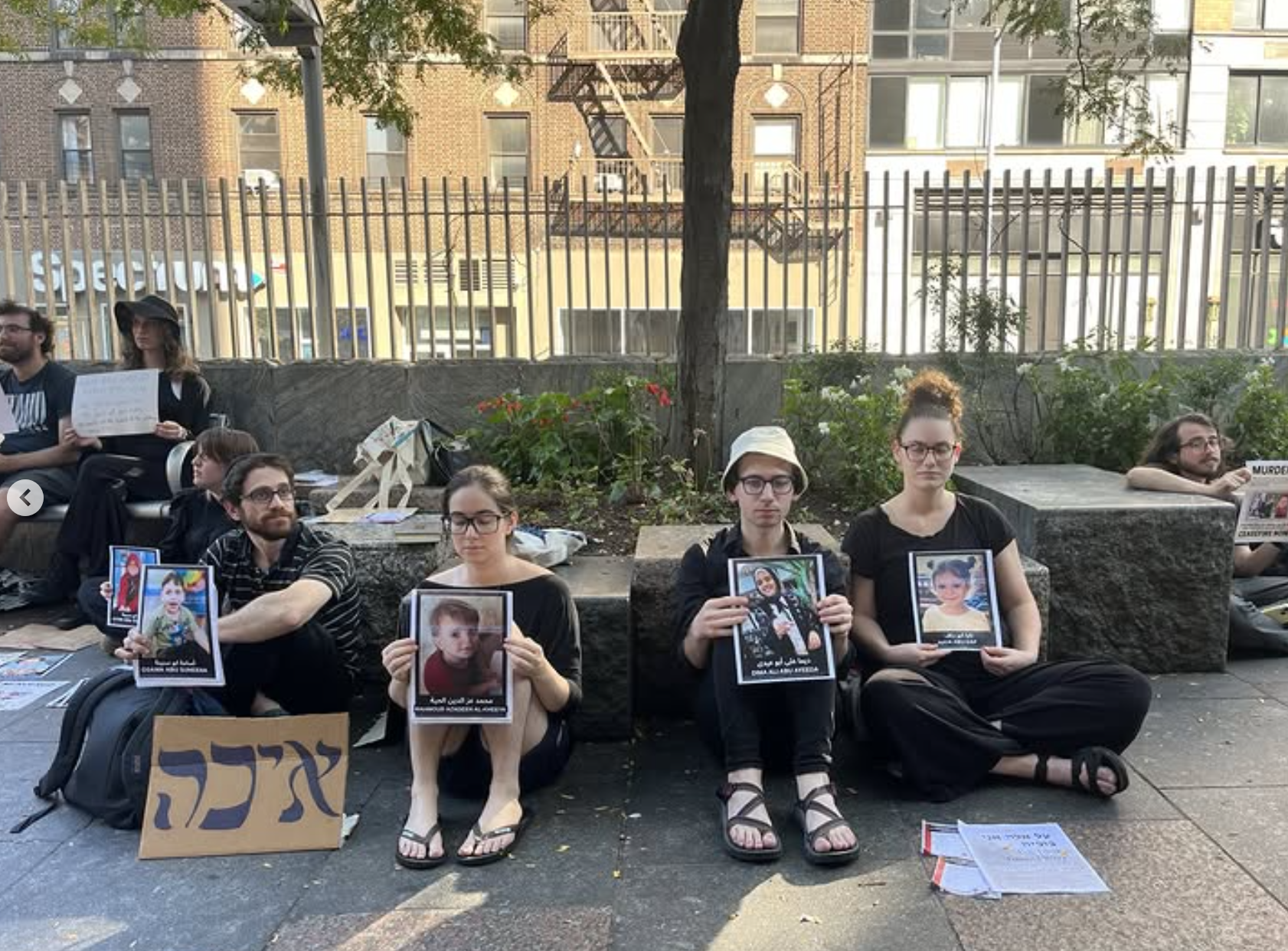 Group of people sitting on the sidewalk in NYC on Tisha B'Av holding photographs of children, with signs and papers around them, participating in a peaceful protest against the Israeli war in Gaza.