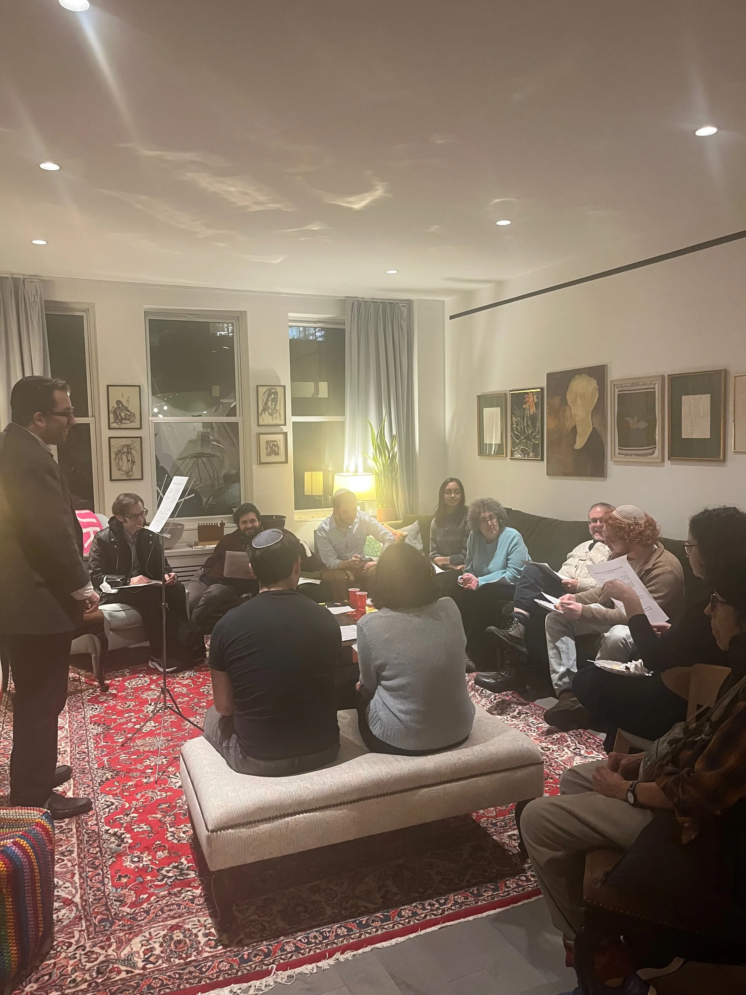 A group of people sitting in a cozy living room arranged in a circle, engaging in a discussion or reading, with a man standing and speaking. The room has framed artwork on the walls, a large window with curtains, a rug with intricate patterns, and a mix of chairs and a bench for seating.