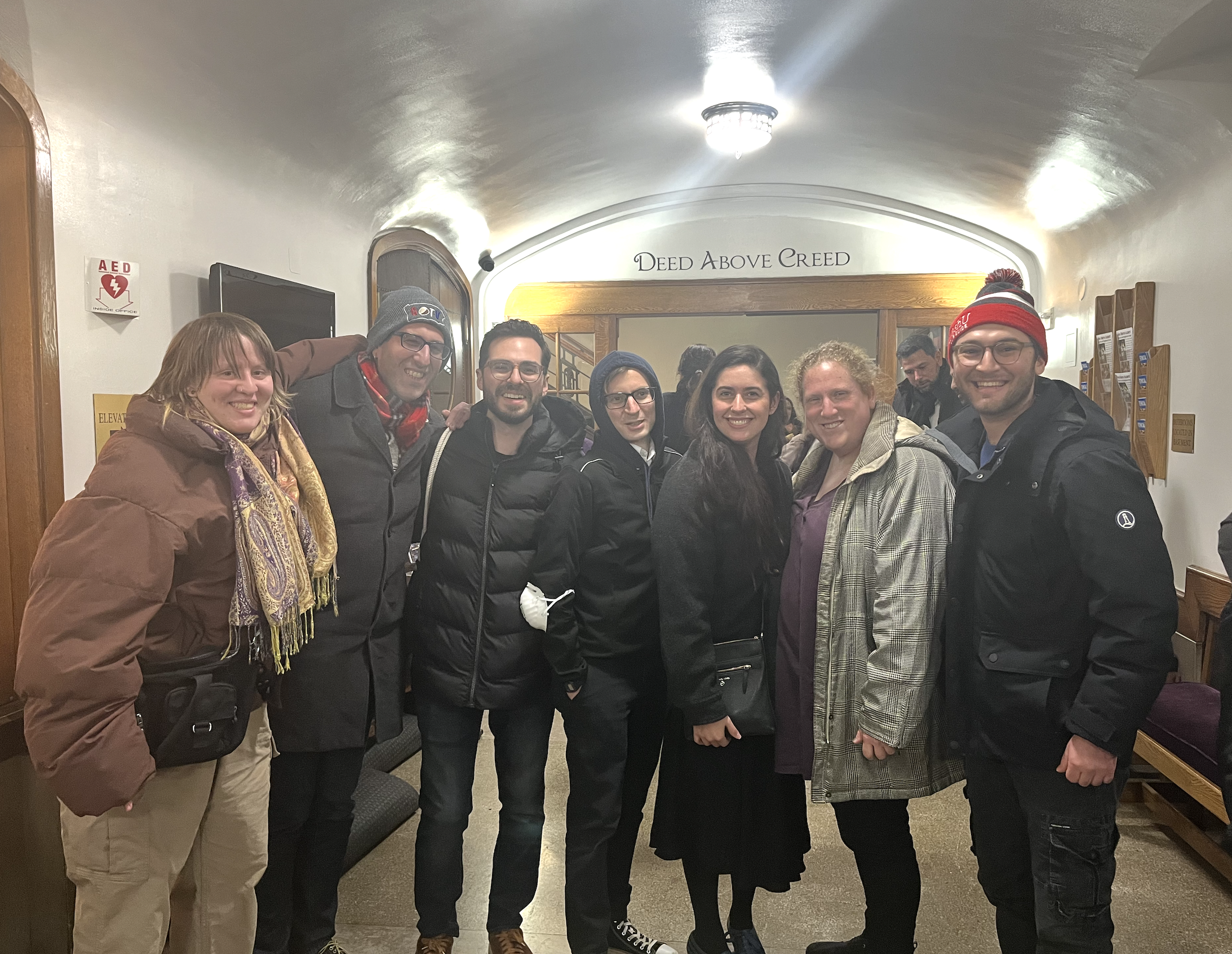 Group of eight people standing together indoors, smiling at the camera, in front of a wooden arch with the words 'Deed Above Creed' written above it.