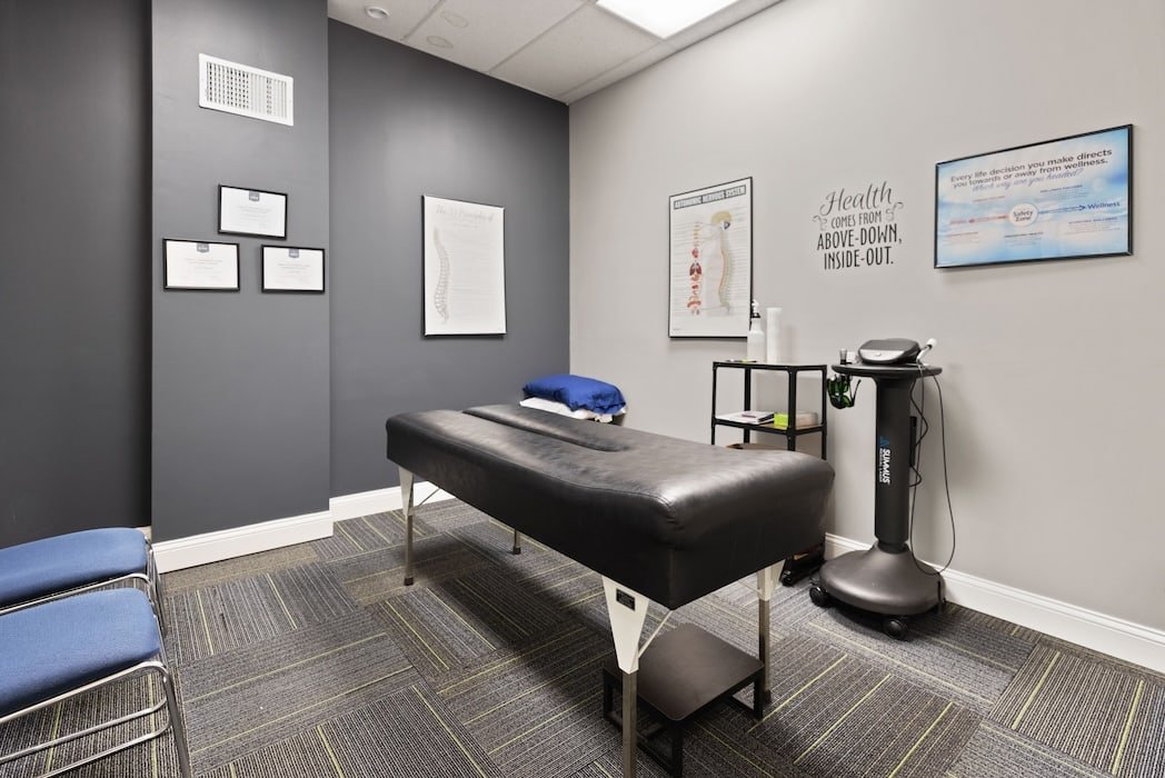 A massage therapy room with a massage table, framed certificates, and medical diagrams on the wall, with a digital screen displaying wellness information.