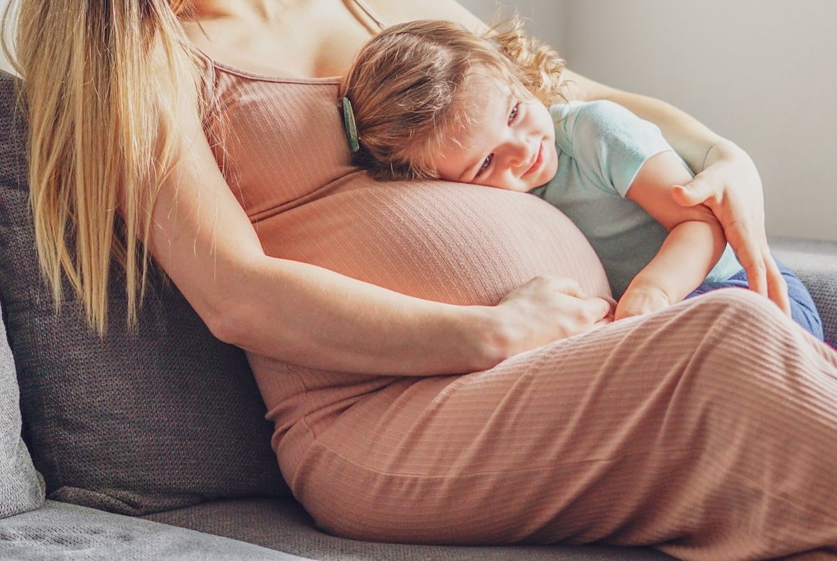 A pregnant woman sitting on a gray couch with a young girl resting her head on her belly.