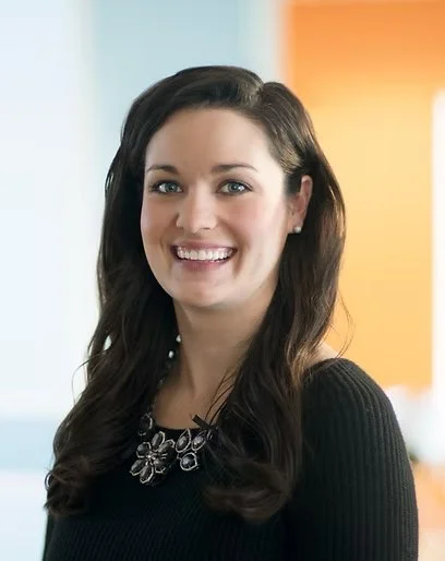 A woman with long dark hair smiling, wearing a black top and a necklace, with a colorful blurred background.