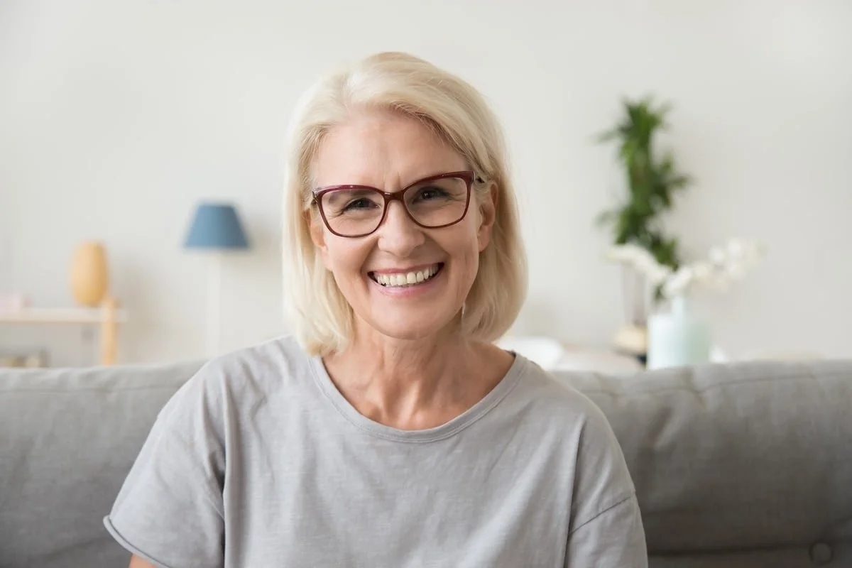 A smiling middle-aged woman with blonde hair and glasses sitting on a grey couch in a bright living room with a plant and a lamp in the background.