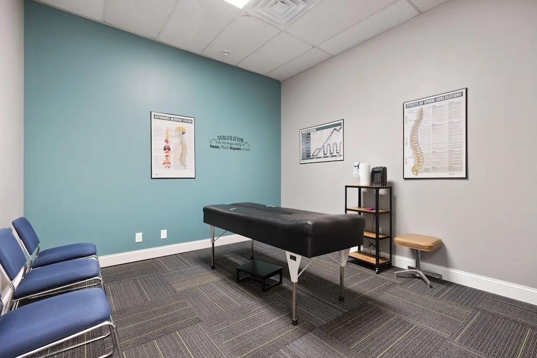 Empty chiropractic examination room with a massage table, three chairs, and posters of spine anatomy on the walls.