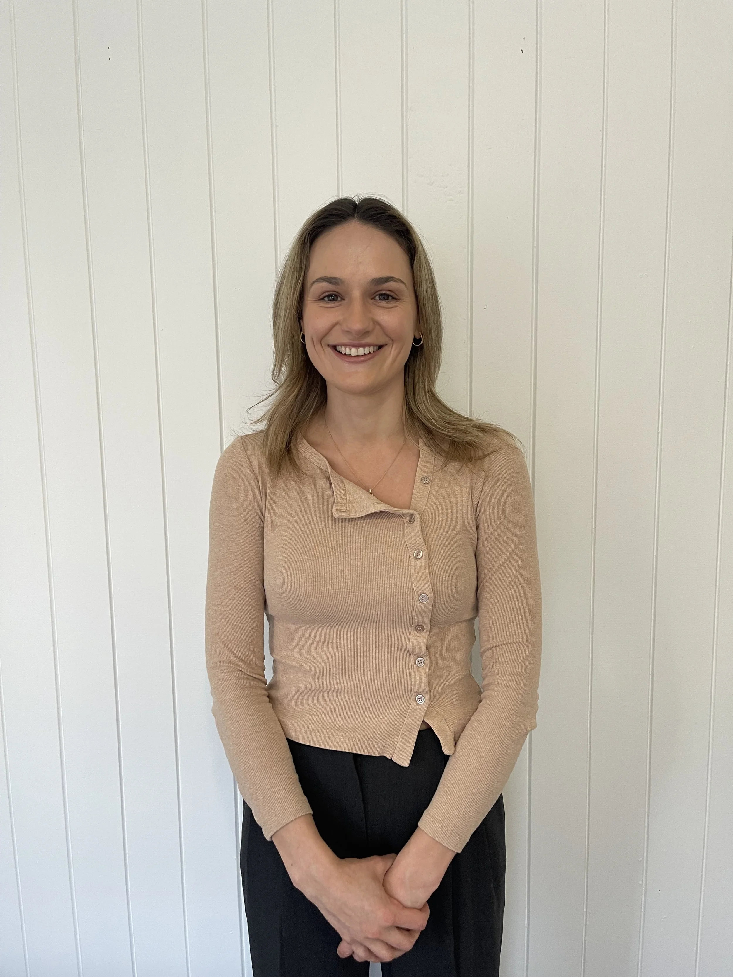 A woman with shoulder-length light brown hair smiling, standing in front of a white paneled wall, wearing a beige button-up sweater and black pants.