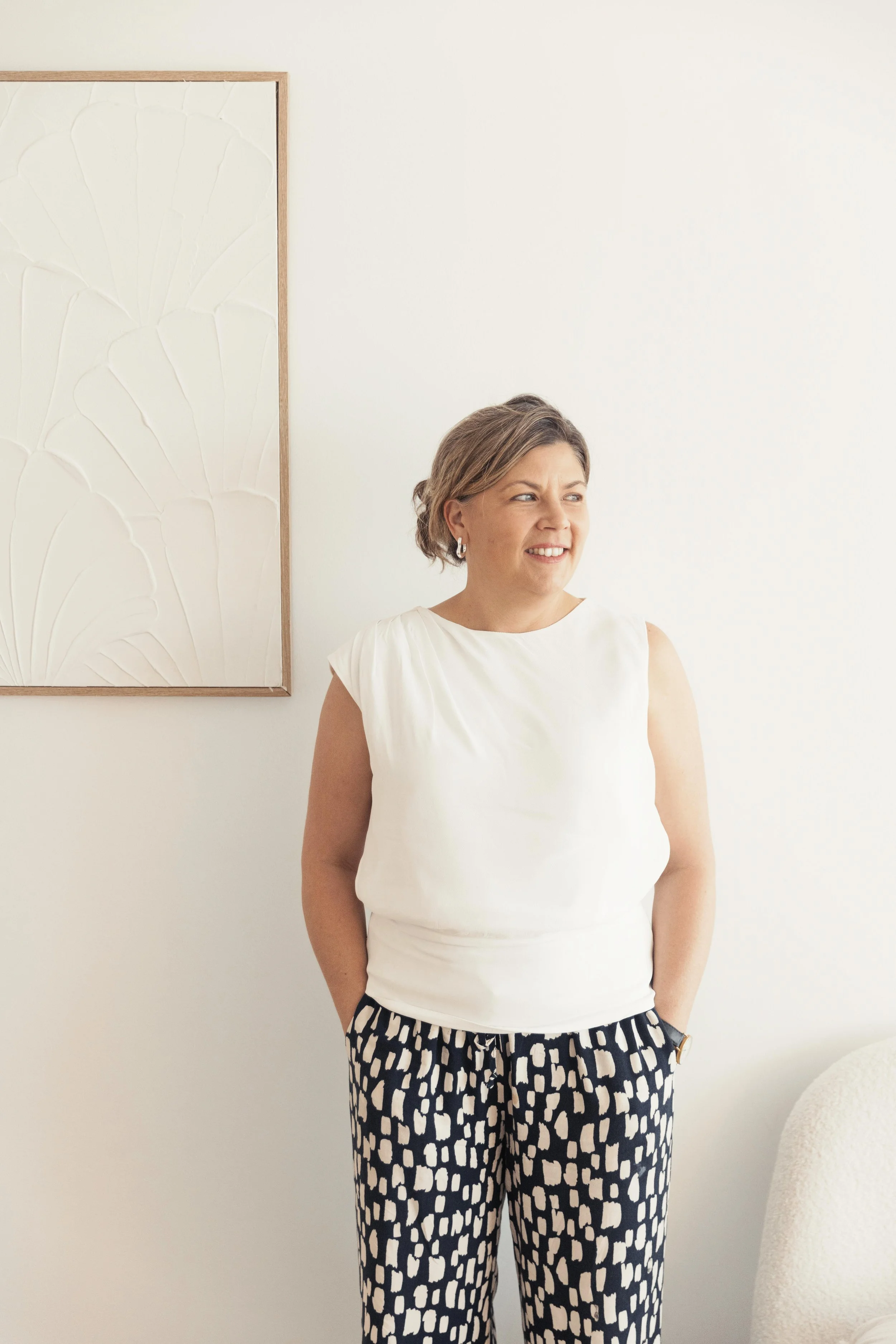 Young woman with long brown hair smiling and crossing her arms, wearing a sleeveless light-colored dress against a plain off-white background.