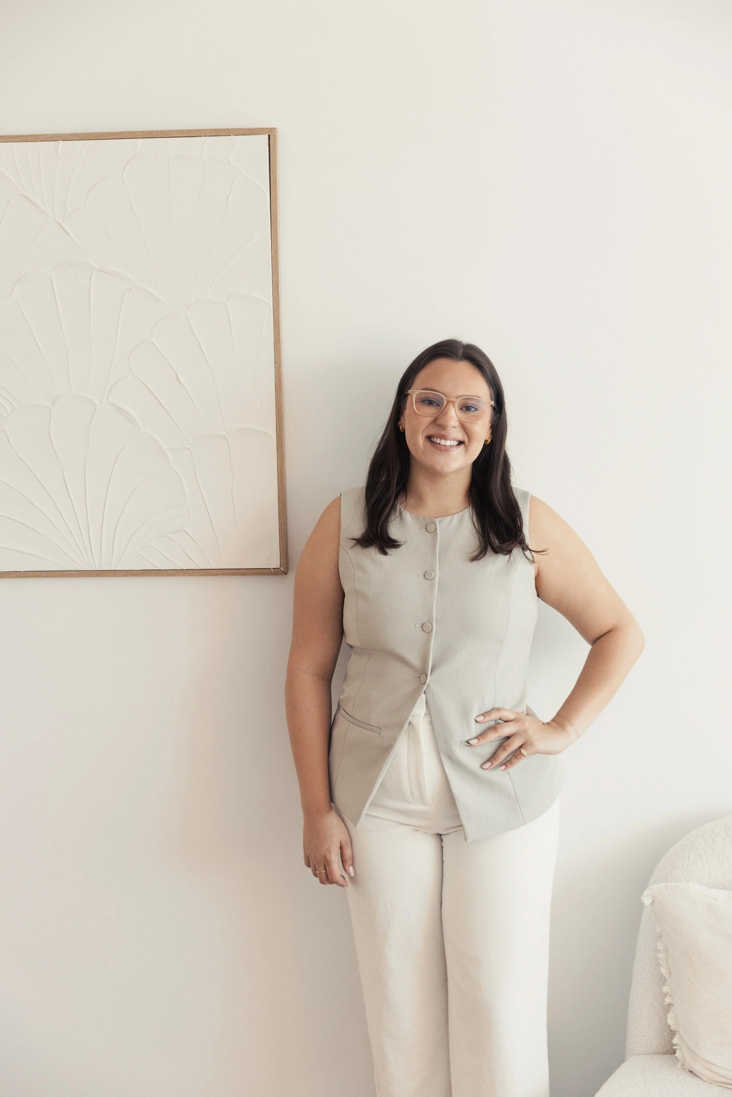 A woman with long blonde hair wearing a light gray top and a white skirt, sitting against a plain beige wall, smiling at the camera.
