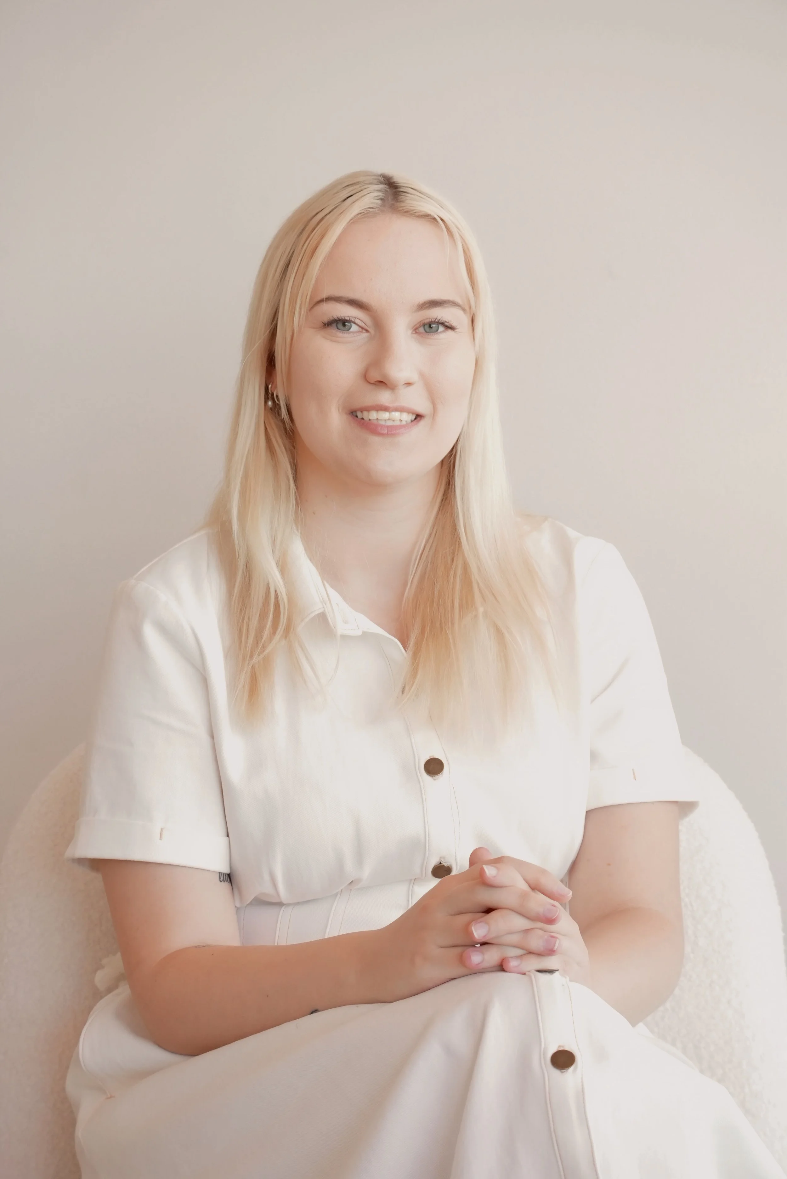 A young woman with blonde hair and blue eyes, wearing a white button-up dress, sitting with hands clasped, smiling against a plain beige background.
