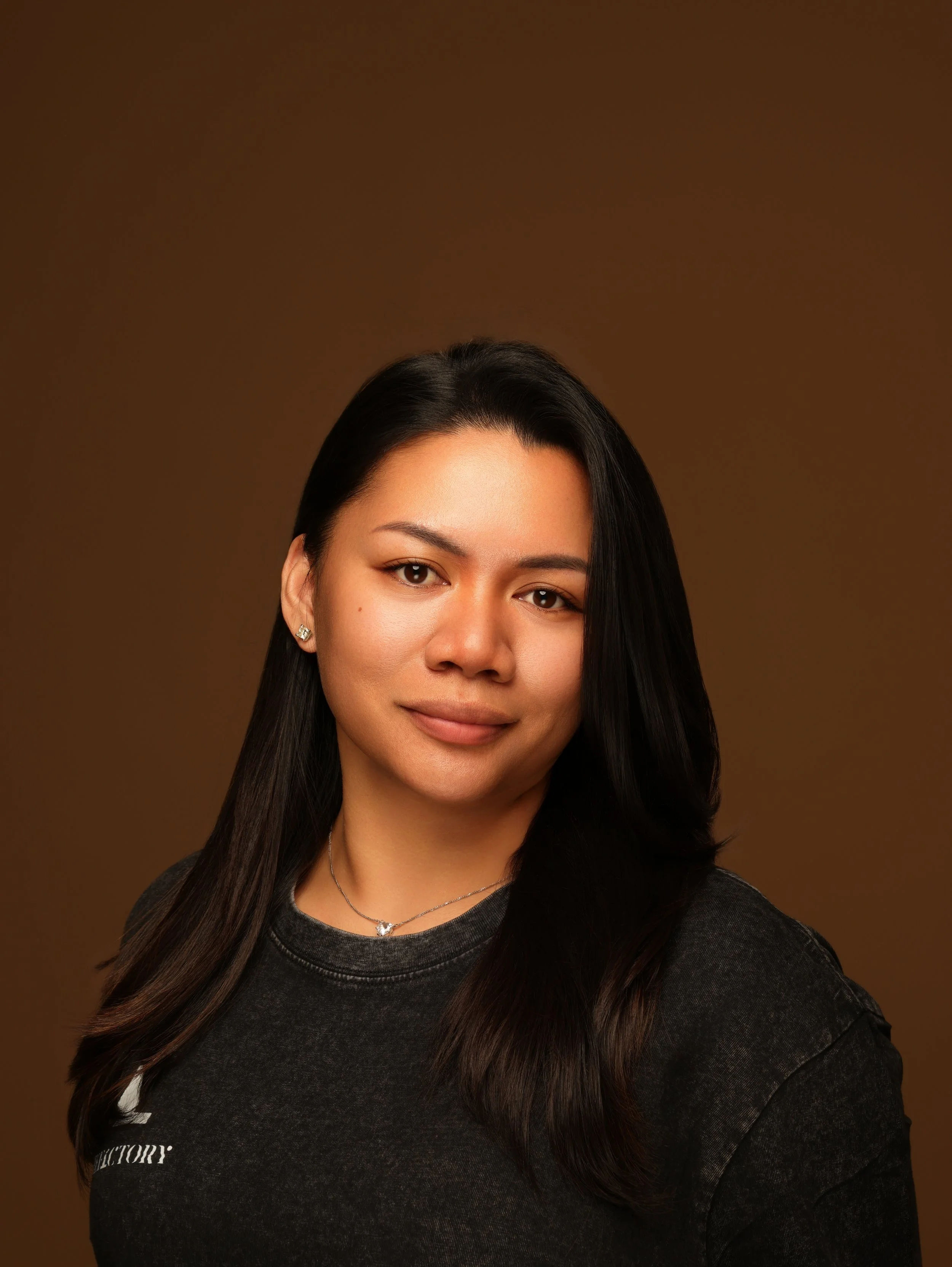 Portrait of a woman with long dark hair, wearing earrings, a necklace, and a dark shirt, against a warm brown background.