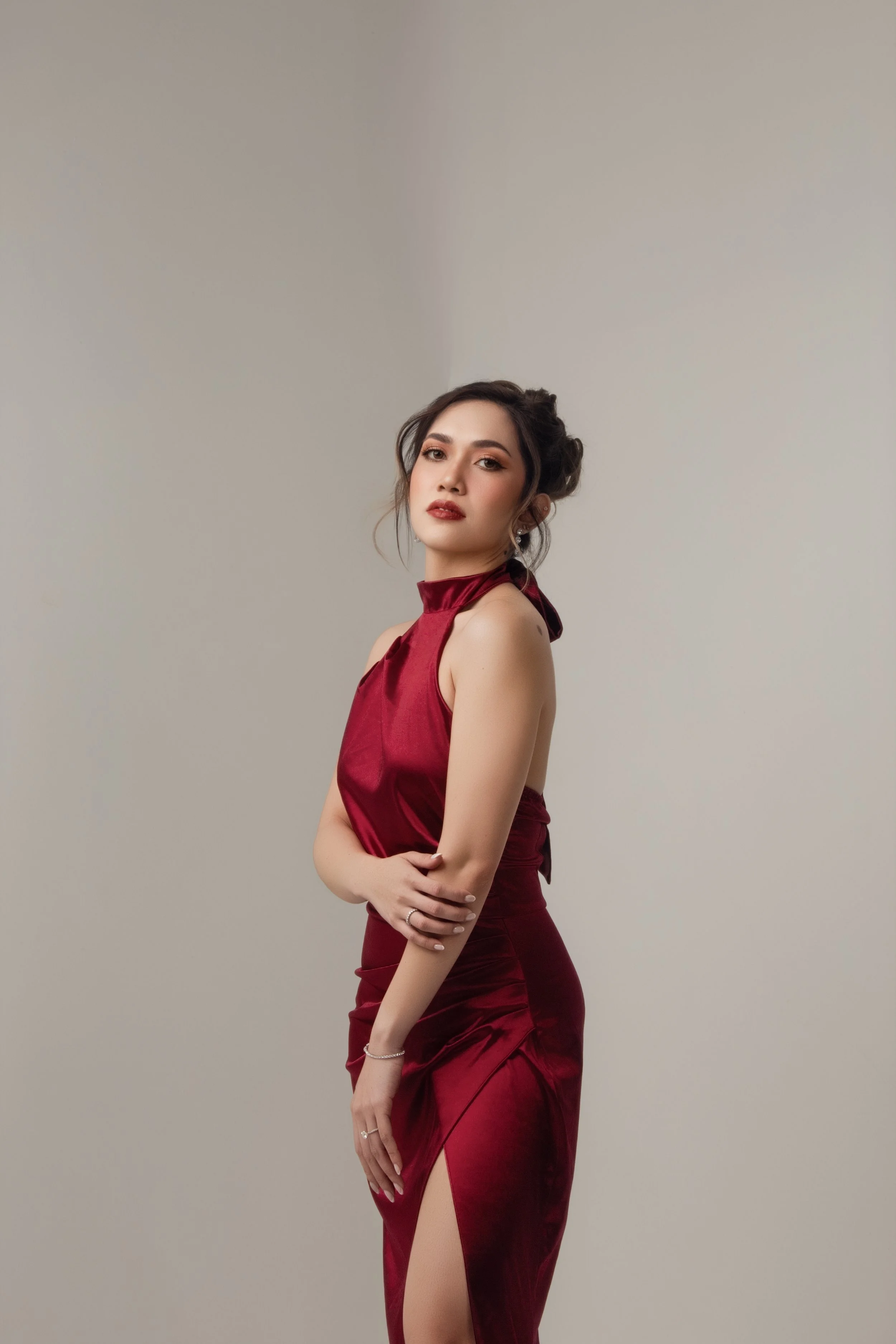 A young woman with dark hair styled in an updo, wearing a sleeveless, red satin dress with a slit, posing against a plain light-colored background.