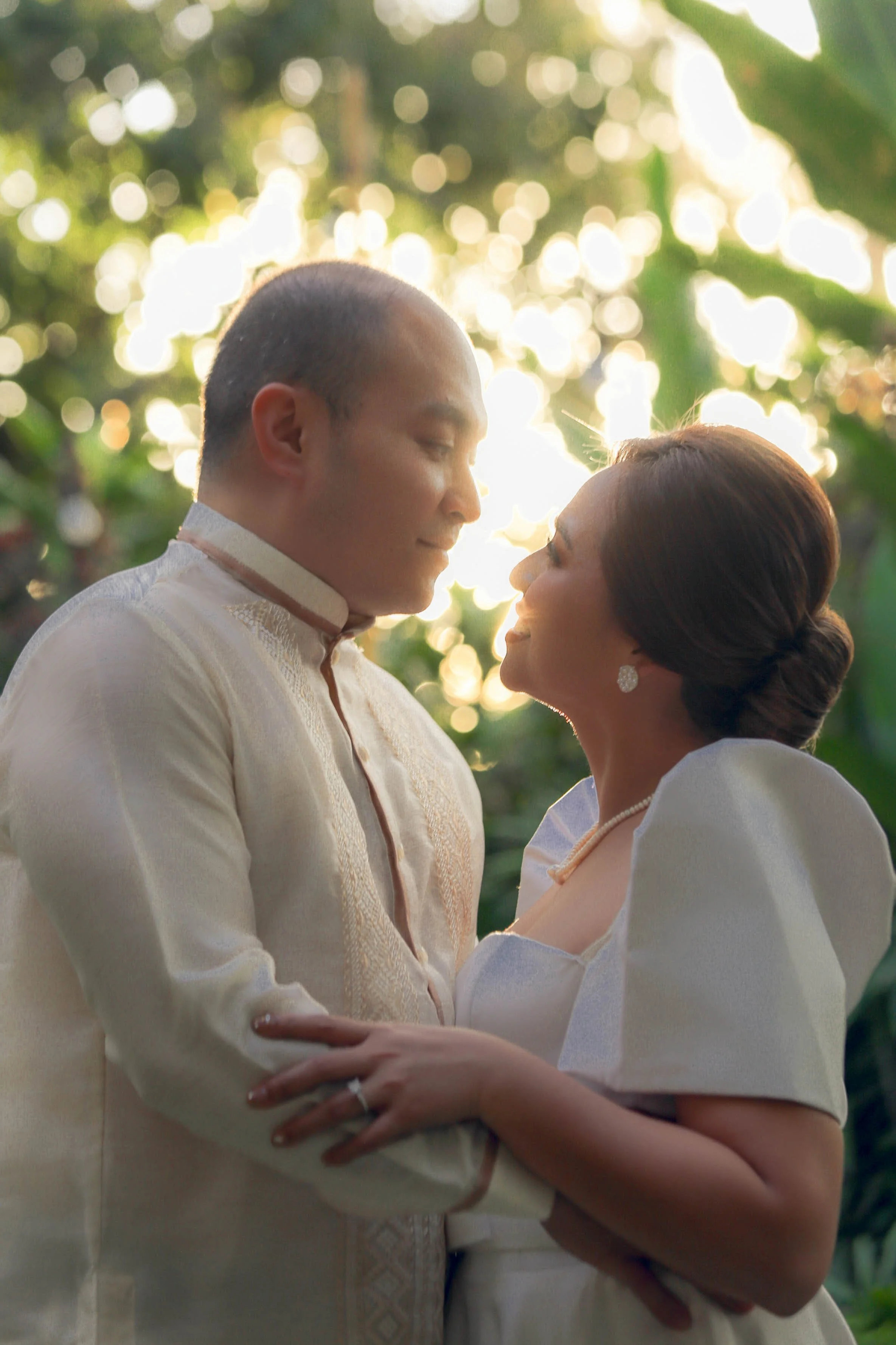 A couple dressed in traditional Filipino barong and formal dress embracing each other outdoors during sunset.