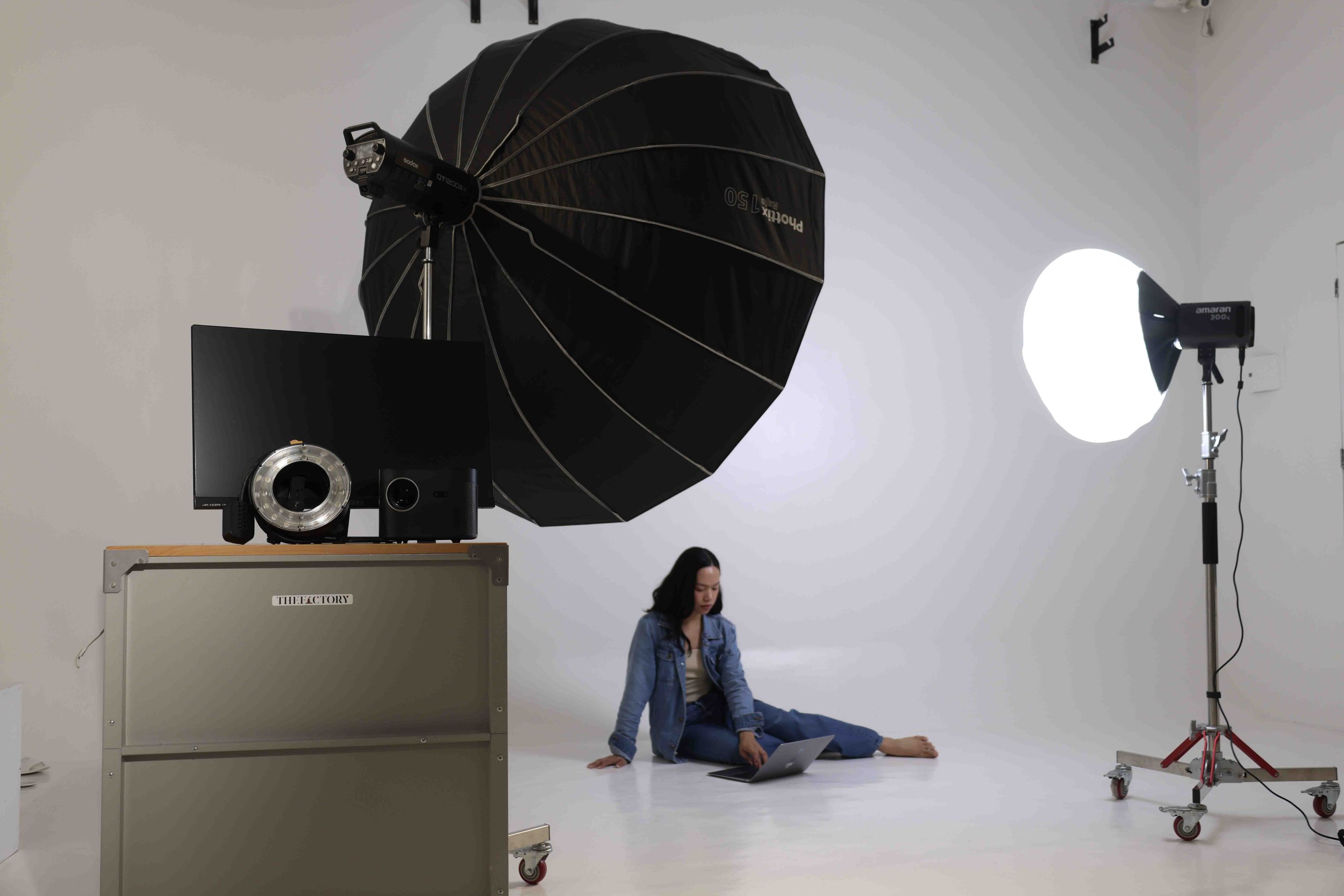 Studio rental setup at The Factory Studio with a white cyclorama, umbrella softbox, continuous light, monitor, and seated model.