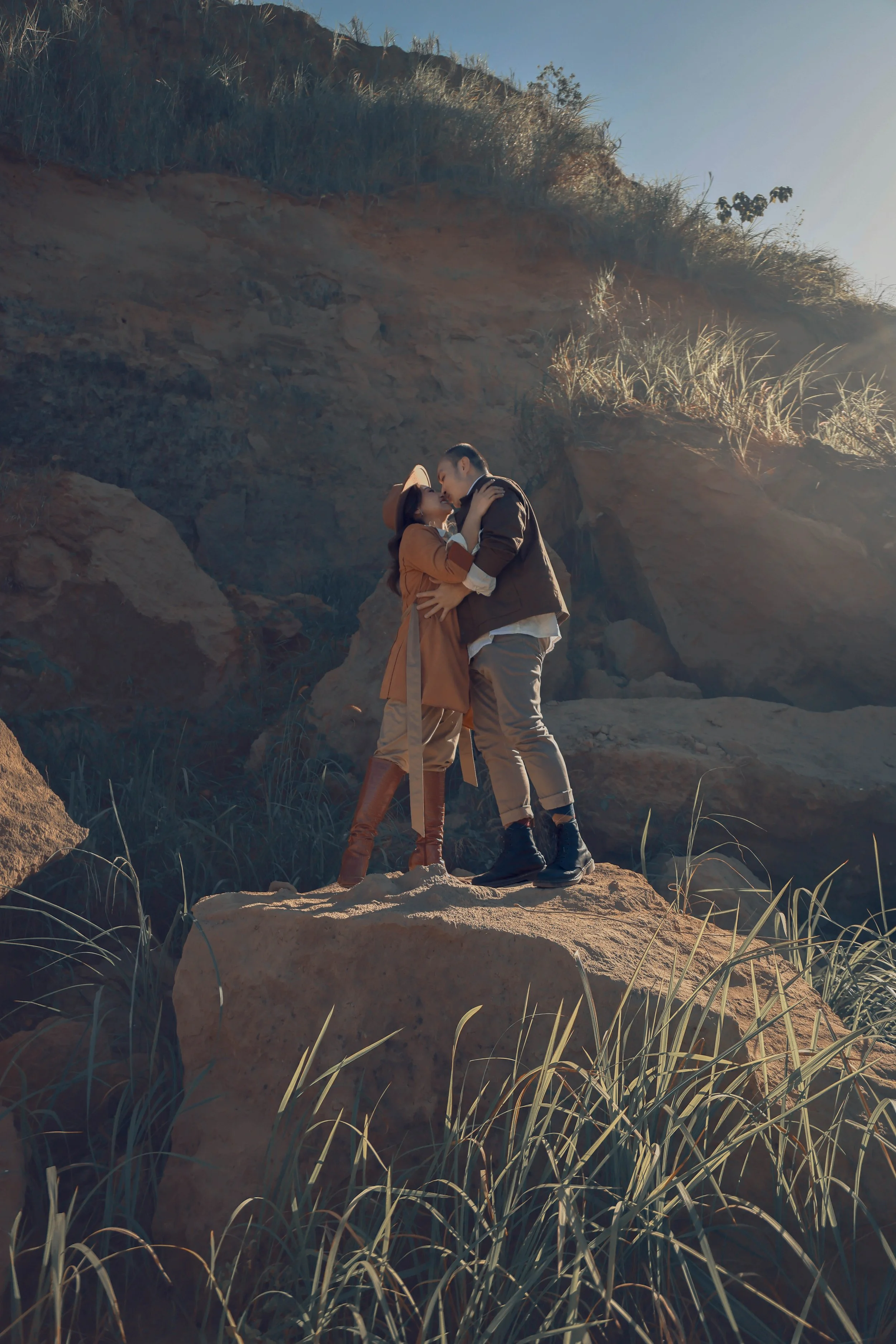A couple embracing on a large rock, surrounded by grass and rocks, with sunlight shining on them from the right.