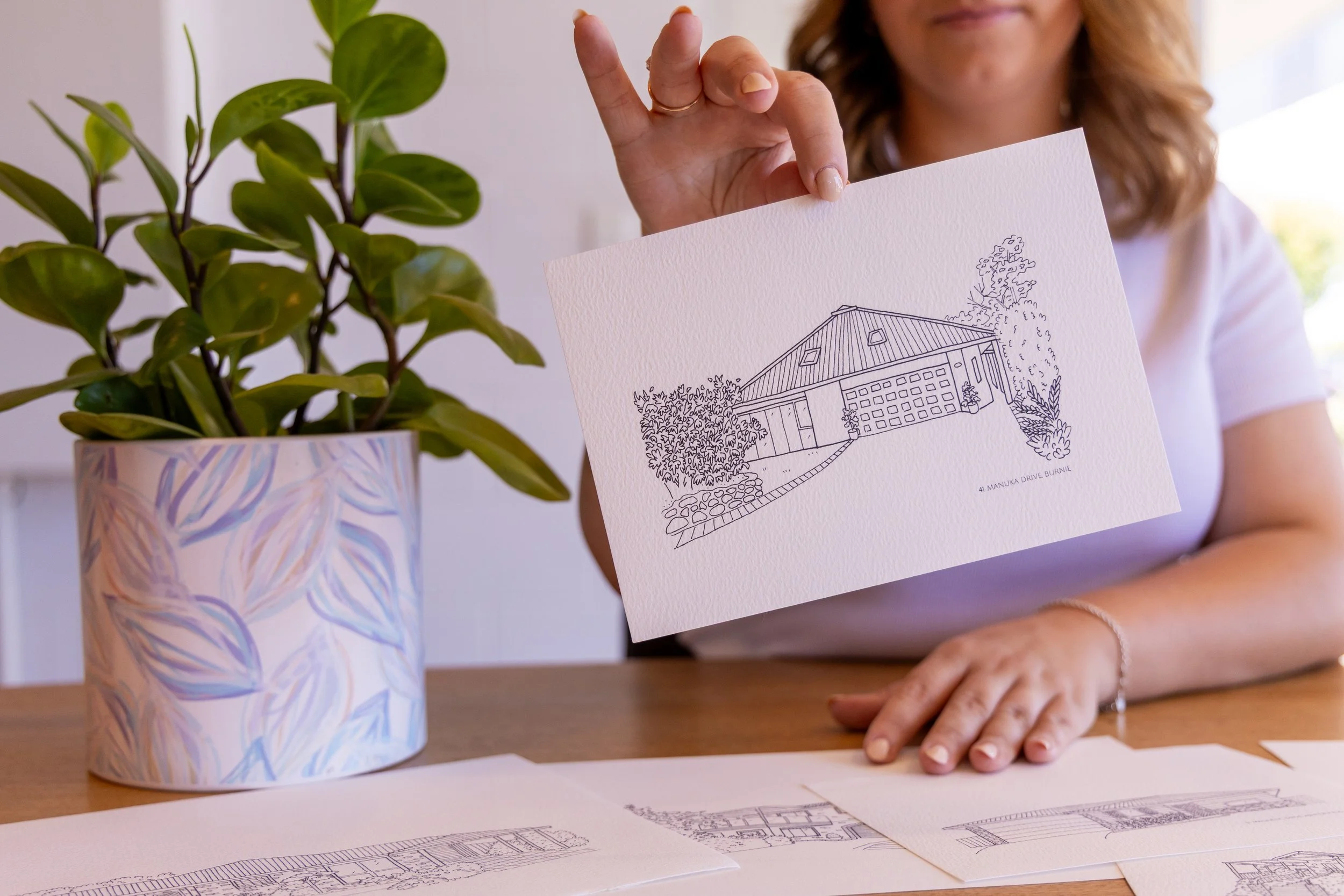Woman holding a piece of paper with a line drawing of a house and trees, sitting at a table with architectural sketches and a potted plant nearby.