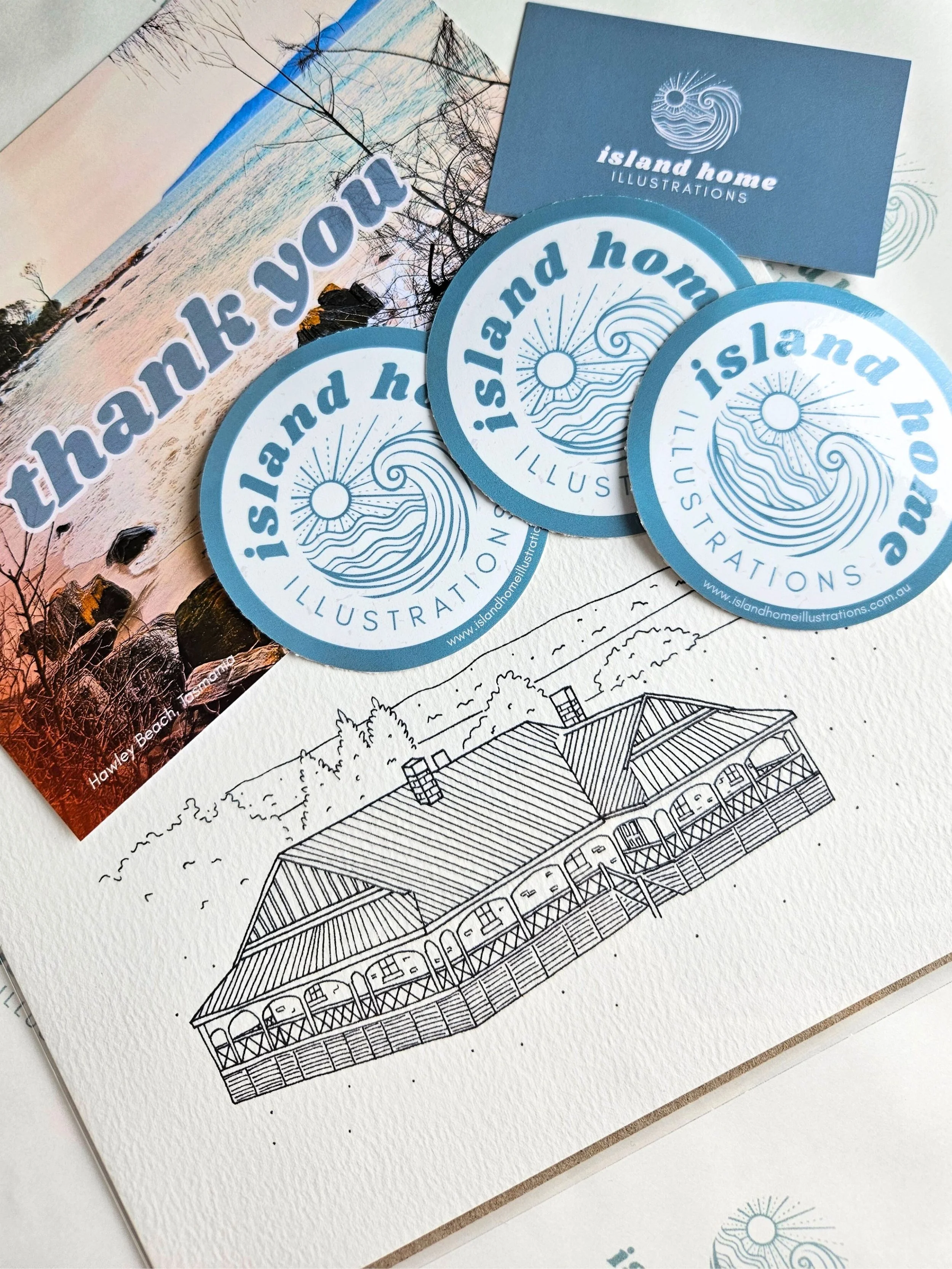 Collection of promotional materials from Island Home Illustrations, including stickers, a business card, an illustration of a seaside house, and a sign with the phrase 'thank you'. 