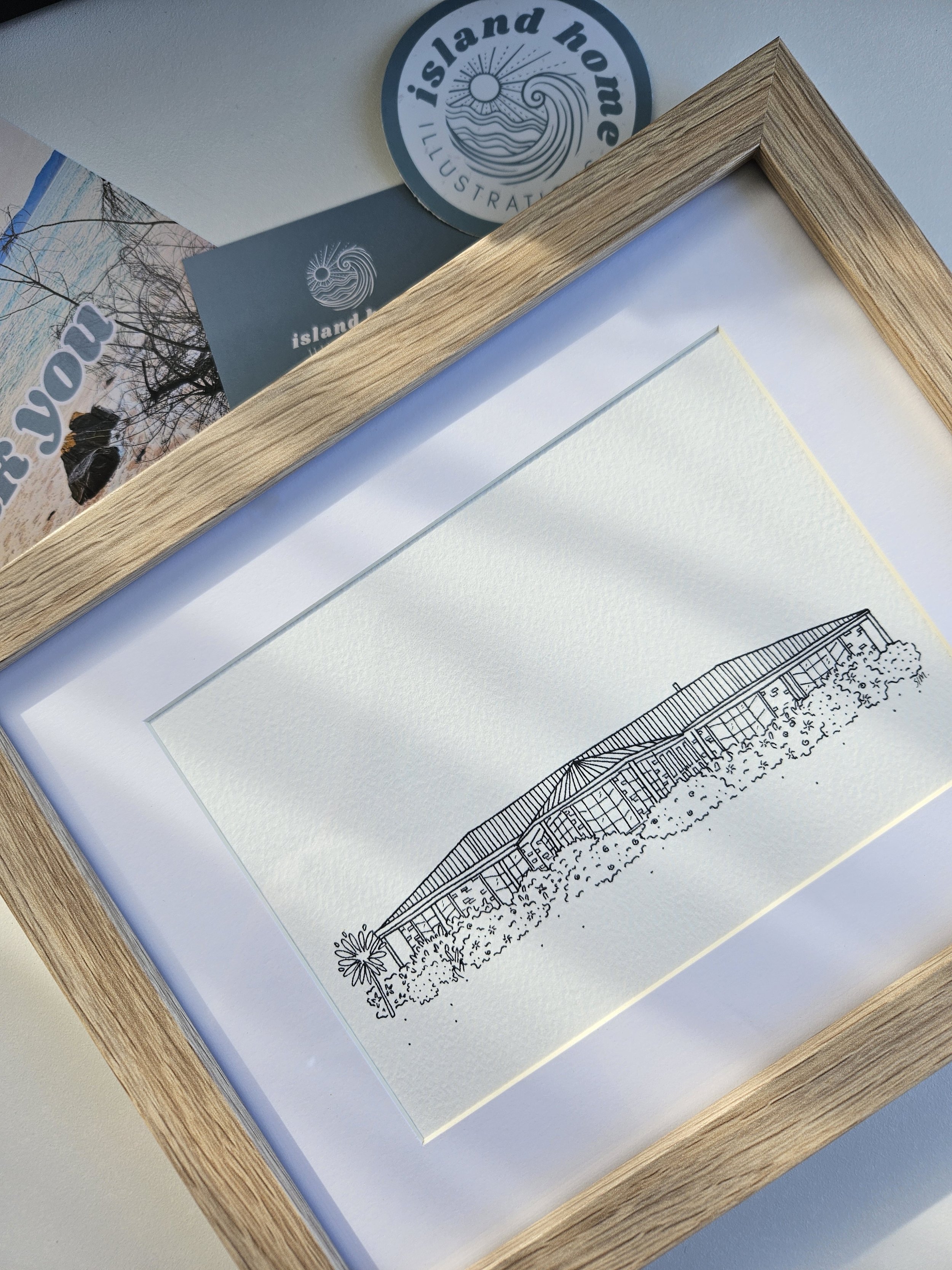 Line drawing of a building with a curved roof, surrounded by bushes and trees, in a wooden frame.
