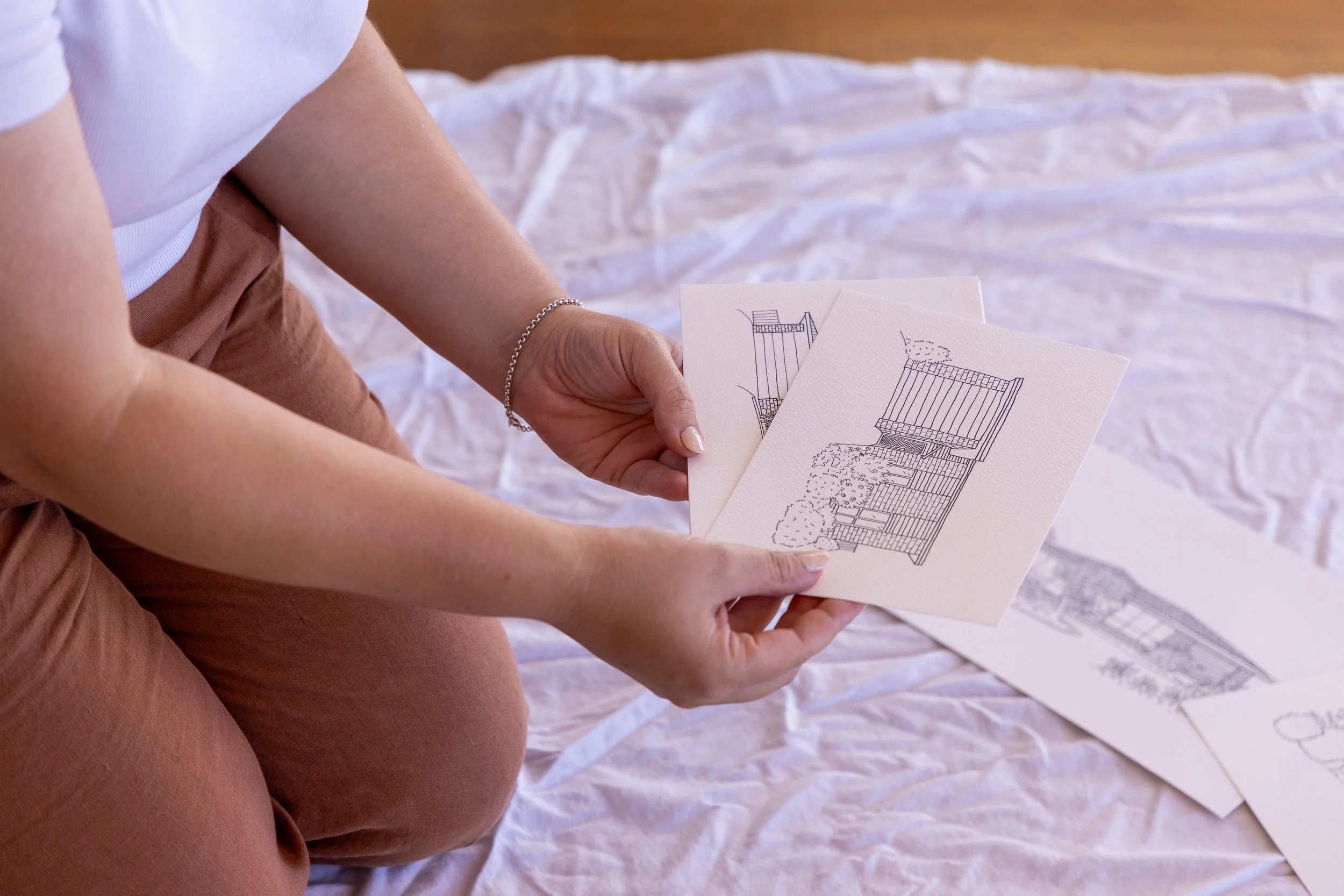 Person sitting on the floor holding two pieces of artwork featuring a house, with additional artwork on the floor.