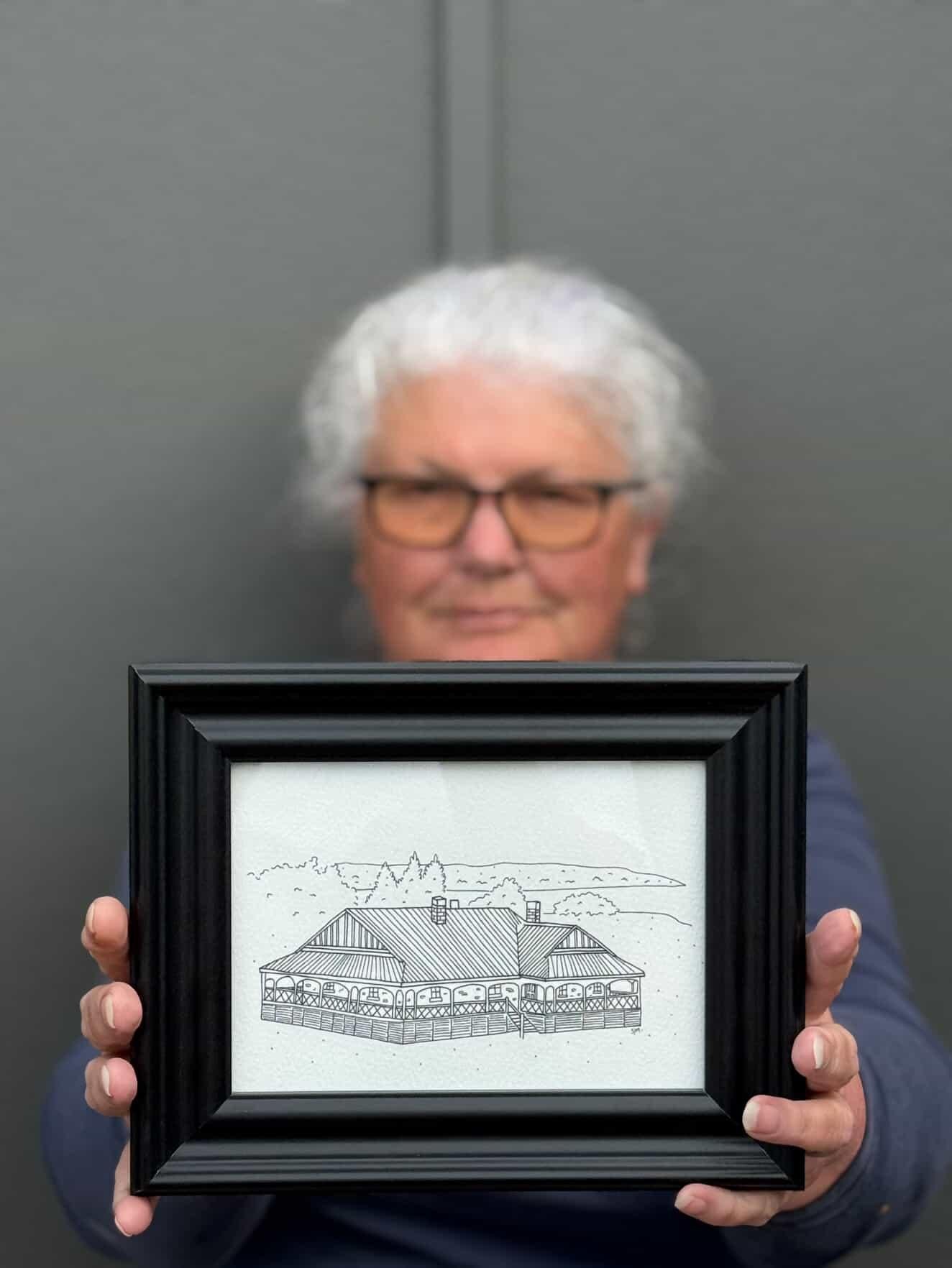 Lady with gray hair and glasses holding a framed illustration of a house with a long porch.