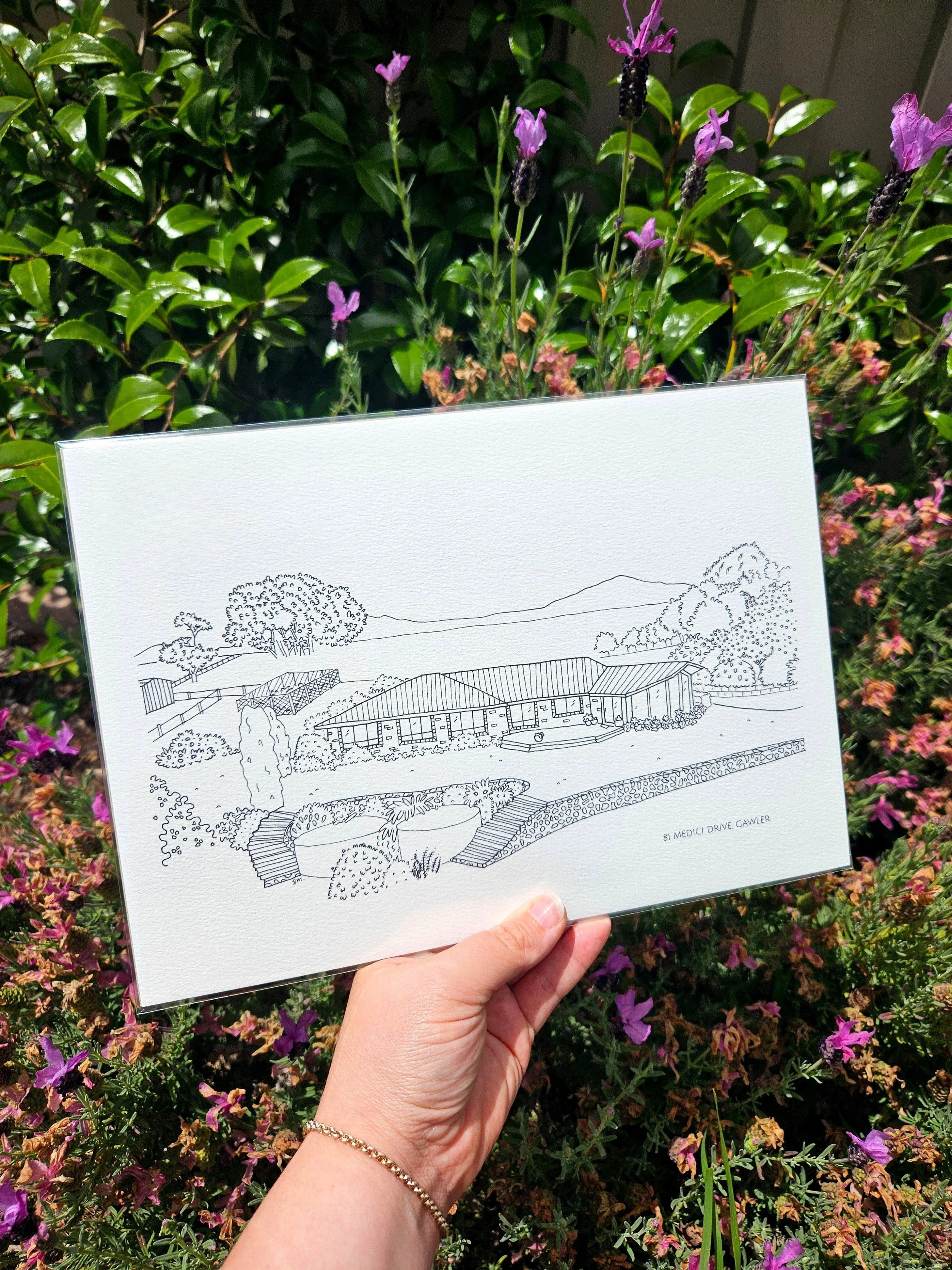 A hand holding a sketch of a house on land with trees, bushes, and a building, against a background of mountains.