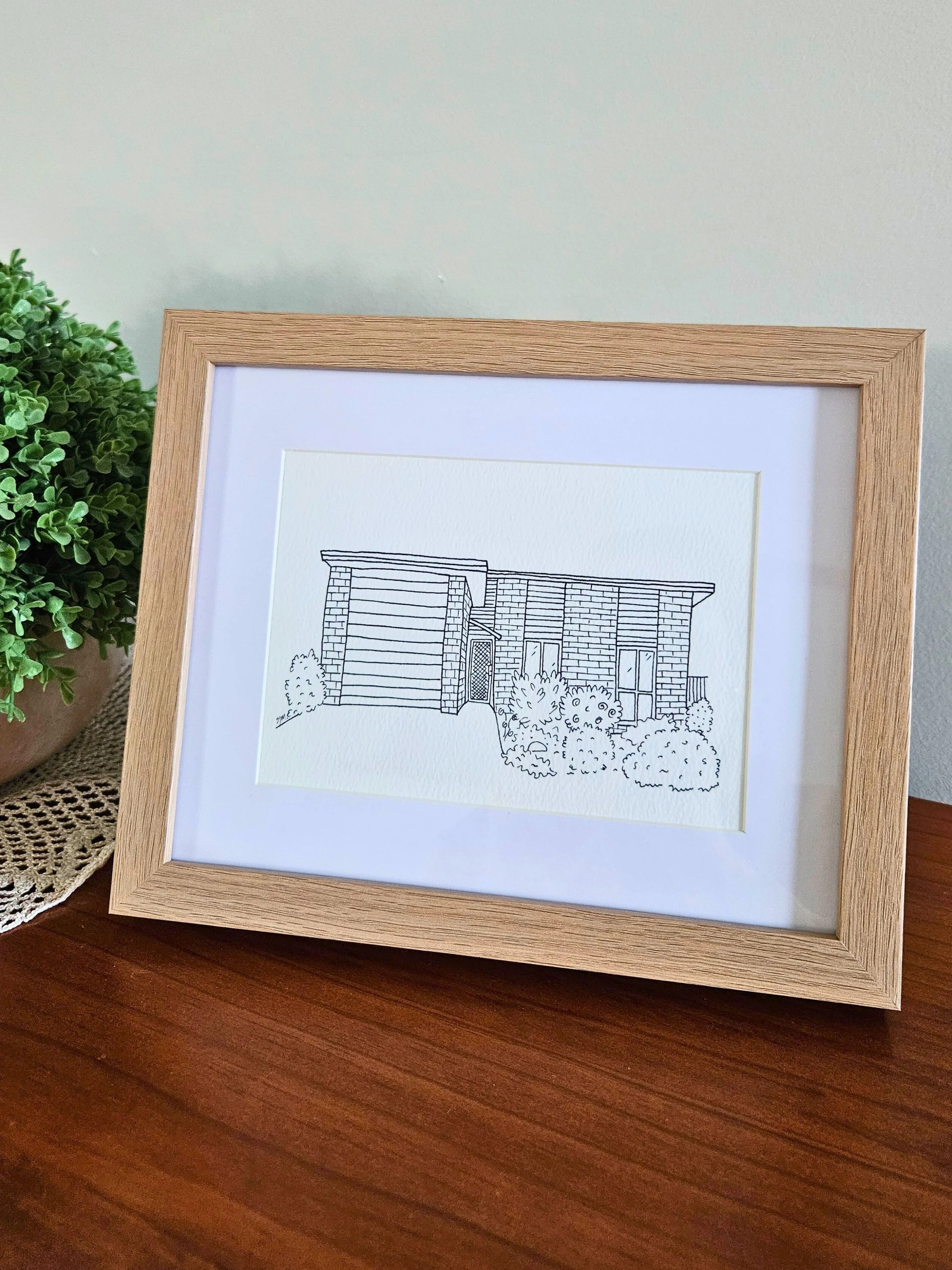 Line drawing of a modern house with bushes and trees in front, framed in a light wood picture frame.