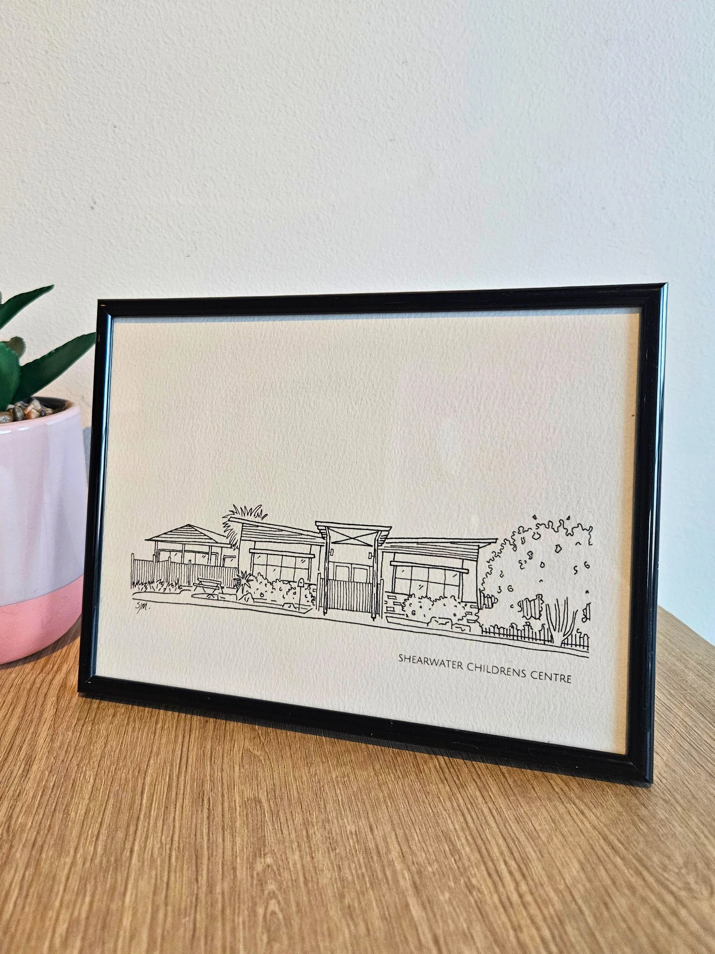 Line drawing of the Shearwater Children's Centre building, framed and leaning against a white wall on a wooden surface, with a potted plant nearby.
