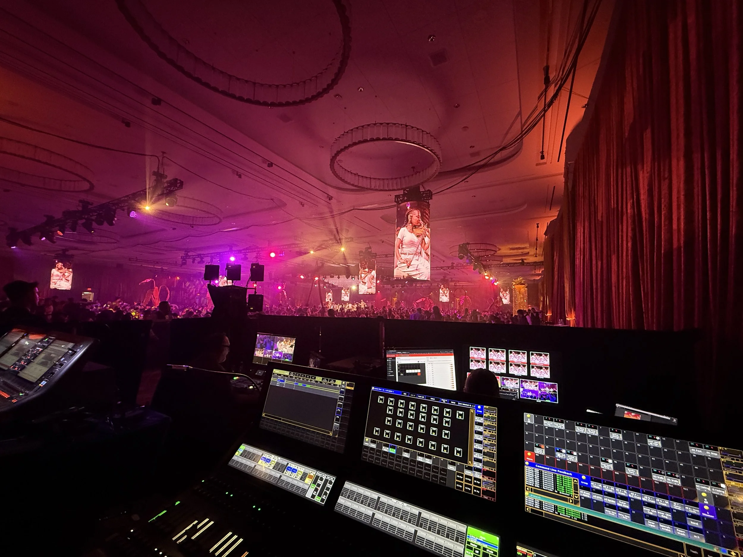Stage with performer singing, large screens displaying the performance, audience in the background, and a control booth with sound and lighting equipment in the foreground.