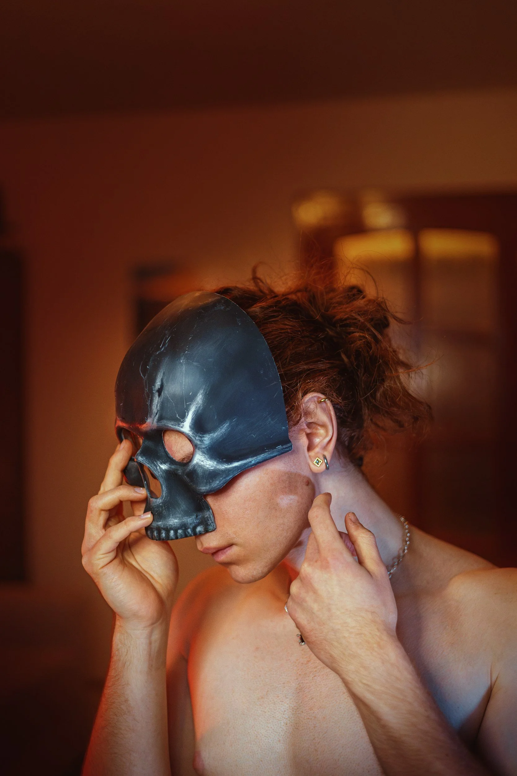 Junge Frau mit roten, lockigen Haaren trägt eine schwarze Totenschädel-Maske, die sie mit der rechten Hand anführt, in einem warm beleuchteten Raum.