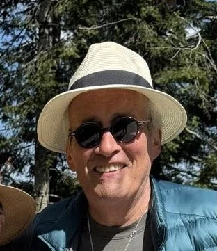 The artist Patrick Walshe wearing a straw hat, sunglasses, and a blue jacket outdoors with trees in the background.
