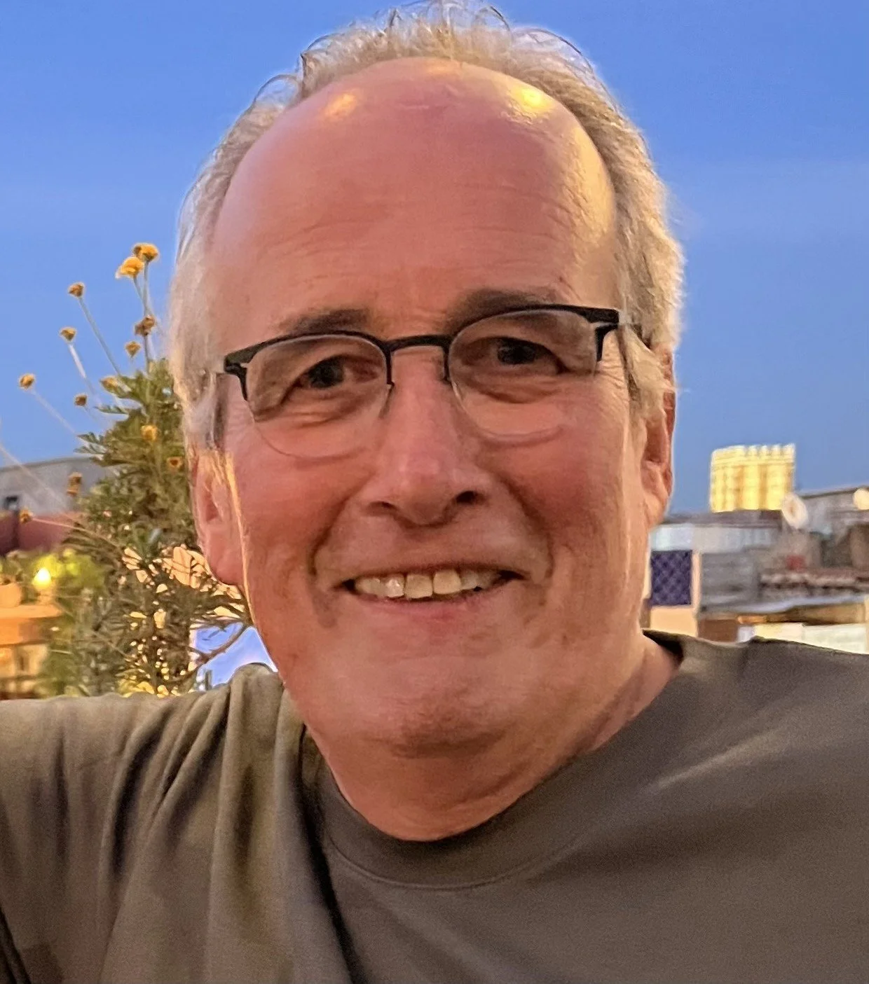 A smiling middle-aged man with glasses, short gray hair, and a beard, taking a selfie outdoors during twilight, with a background of a rooftop with plants and city buildings.