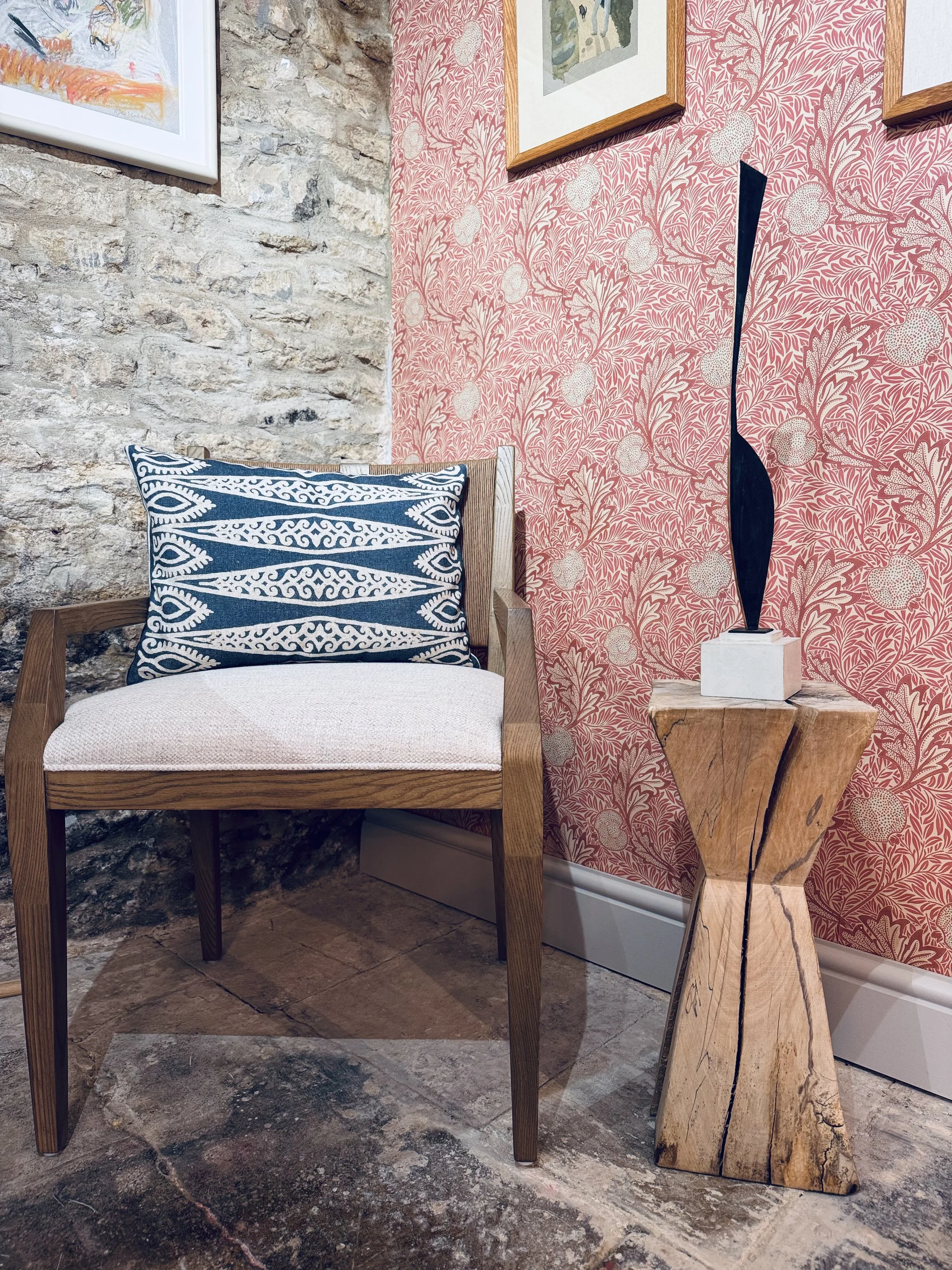 A wooden chair with a blue and white patterned pillow, next to a carved wooden side table with a black sculptural object, set against a pink wallpaper with a fruit and leaf pattern, and an exposed stone wall.
