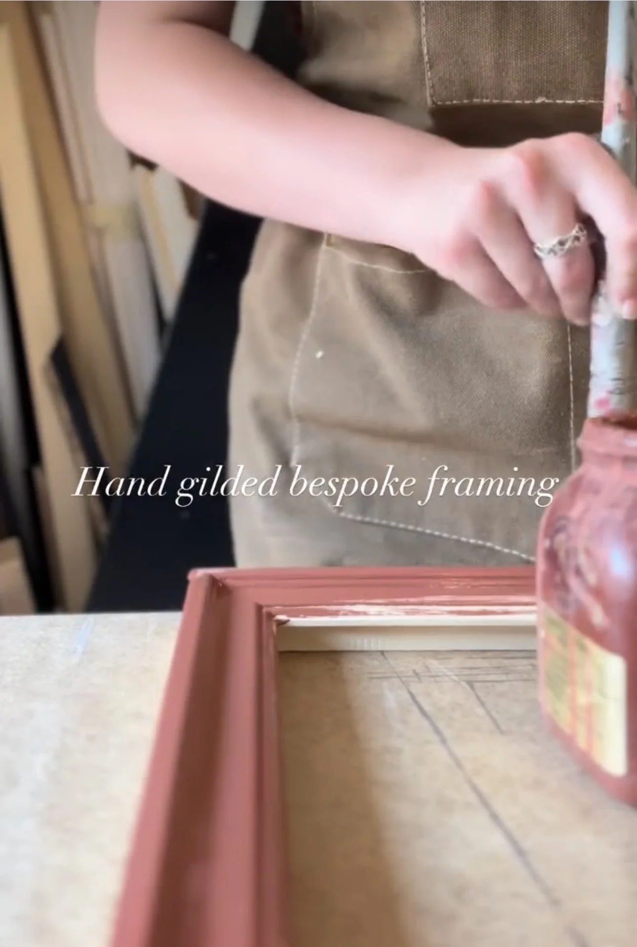 Close-up of a hand with a silver ring, resting on a pink tray surface. Background shows a person's arm, a brown fabric surface, and a mirror reflecting the scene with a caption that reads 'Hand gilded bespoke framing.'