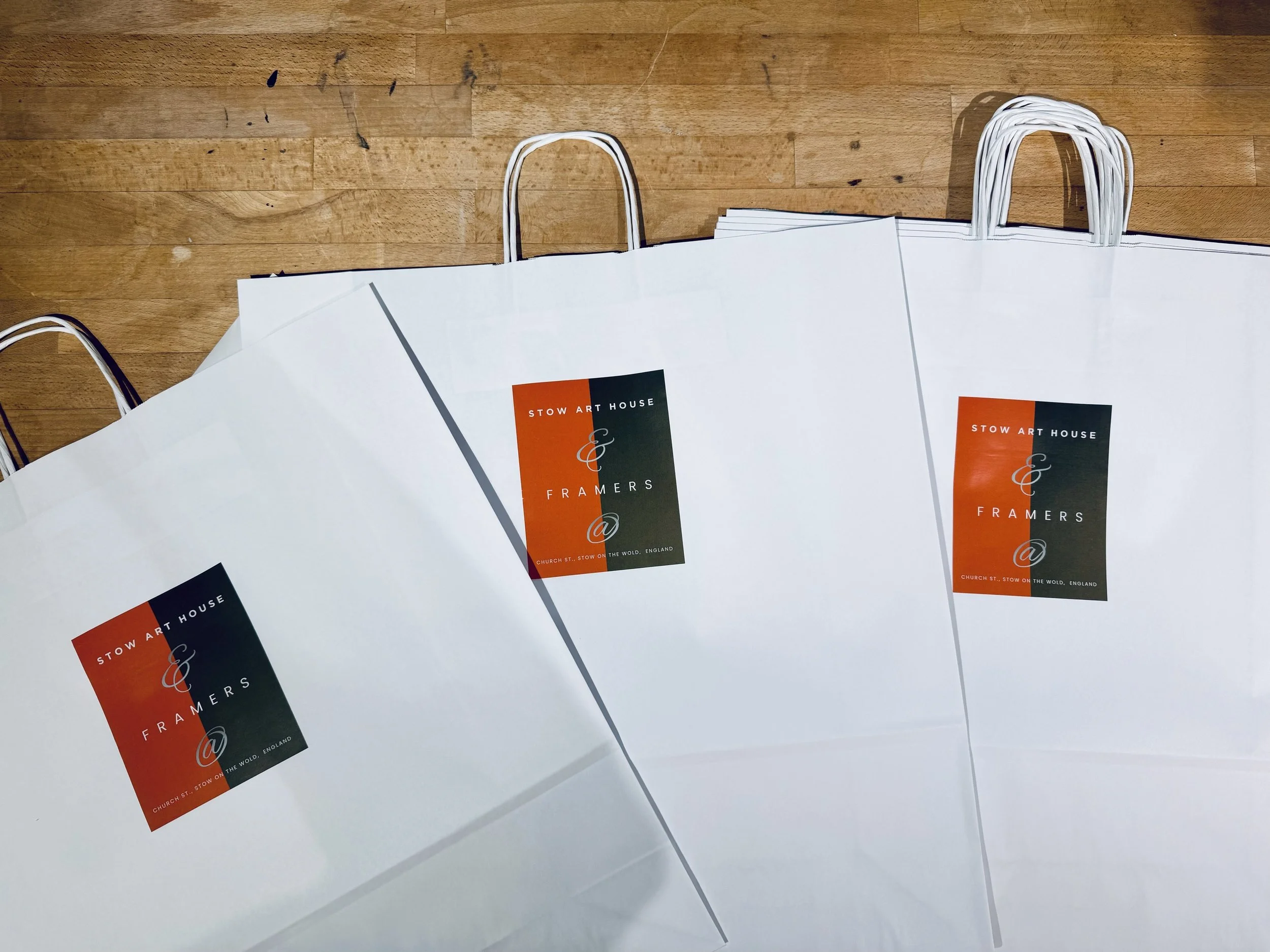 White paper shopping bags with orange and black logo placed on a wooden floor, some handles visible and overlapping.
