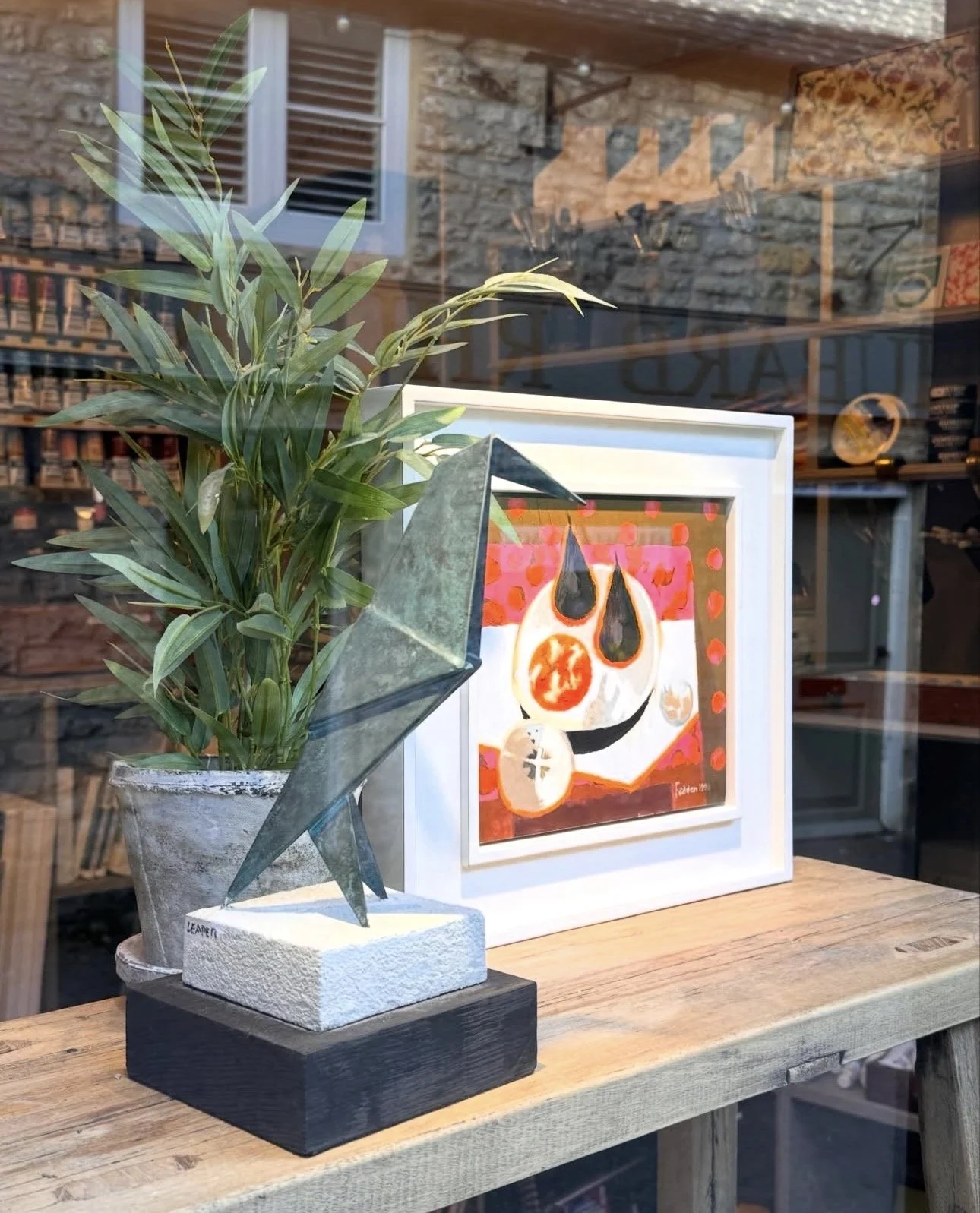 A potted green plant and an abstract art painting with orange, black, white, and pink colors displayed inside a store window, with reflections of the outside environment visible on the glass.