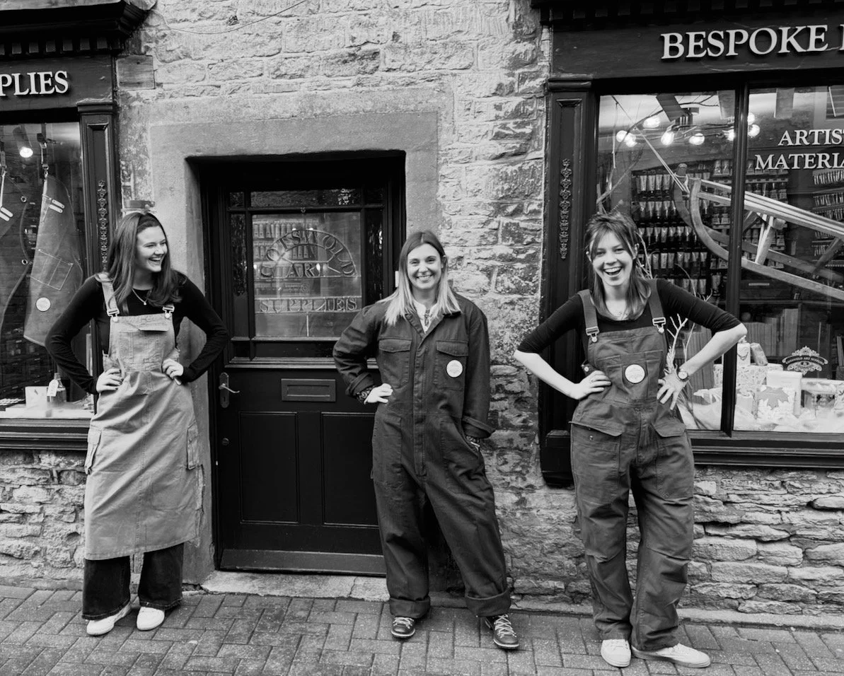 Three women standing outside a shop, smiling and posing for the camera, in front of a stone wall and window with art supplies and books inside.